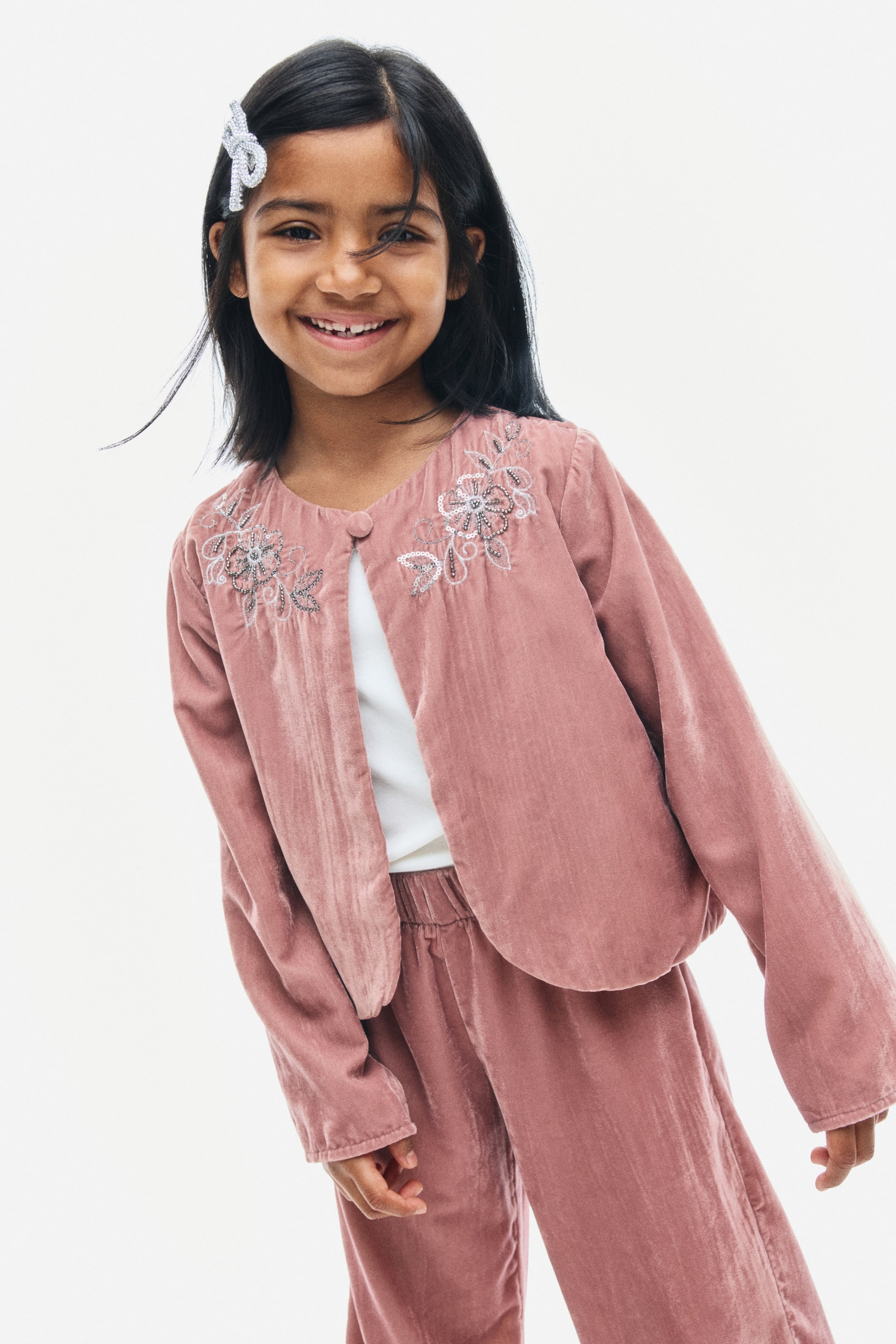 View larger image: A dusty pink crinkled velvet jacket is facing the front, showcasing silver floral bead embroidery on the upper chest and a round neckline with a fastened covered button. The long-sleeved jacket is draping open below the single-button closure over a white top and matching velvet trousers.