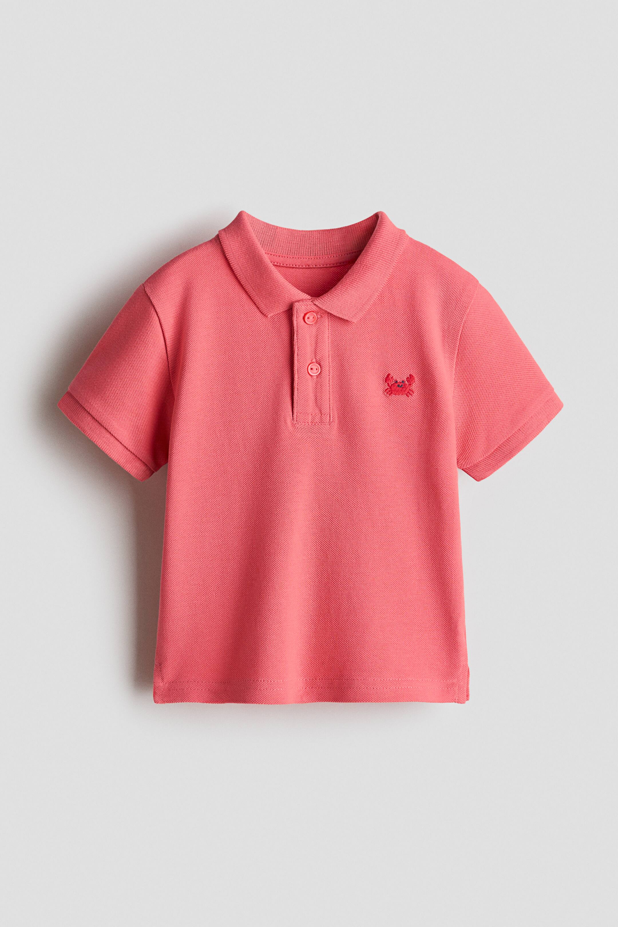 View larger image: A coral short-sleeved polo shirt shown front-on, with a ribbed collar, a two-button placket with matching buttons and ribbed cuffs. It displays a piqué knit texture and a small embroidered crab on the left chest.