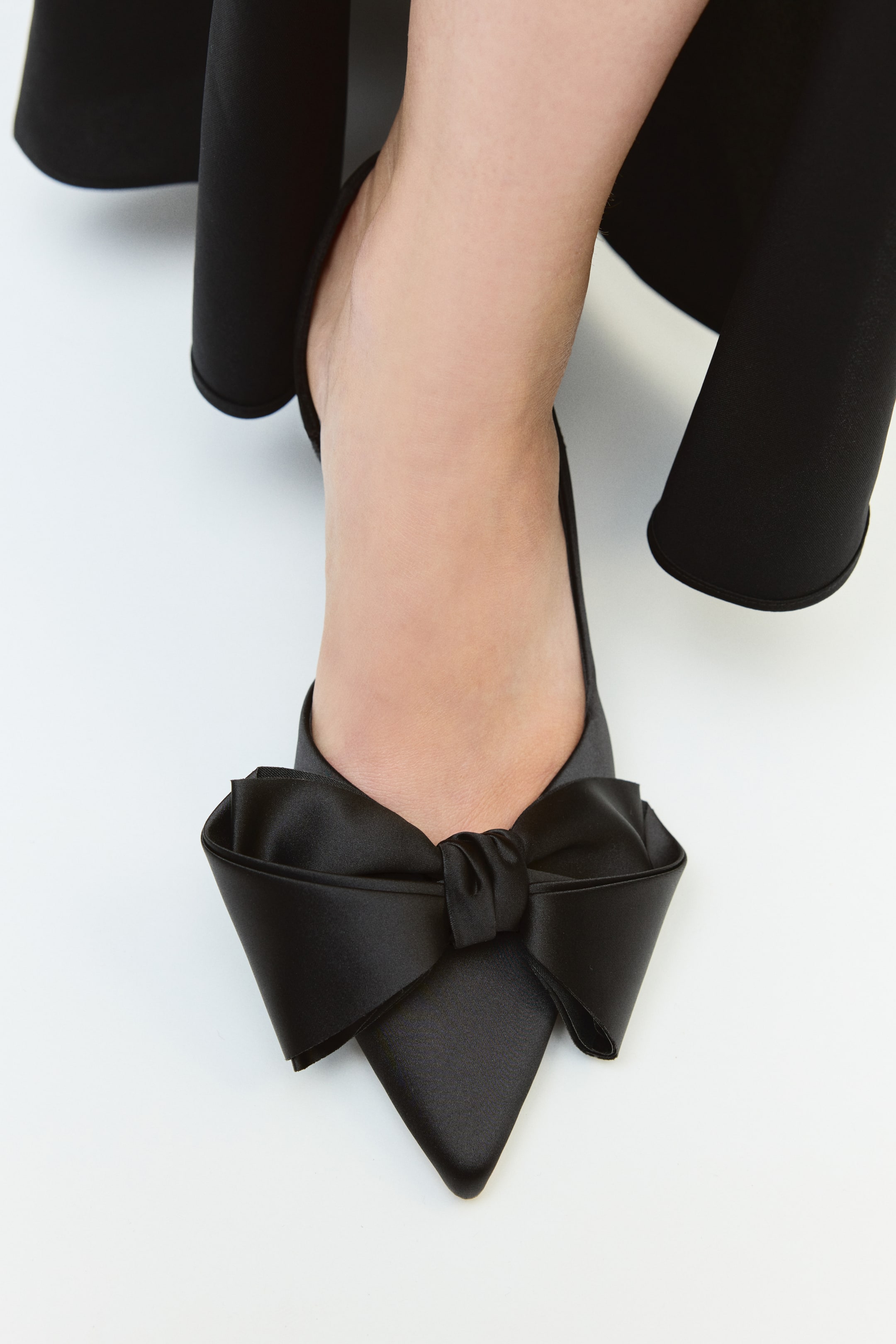 View larger image: A black satin shoe with a pointed toe and a large decorative bow at the front is being worn by the model. The shoe is styled with a draped black garment visible around the leg.