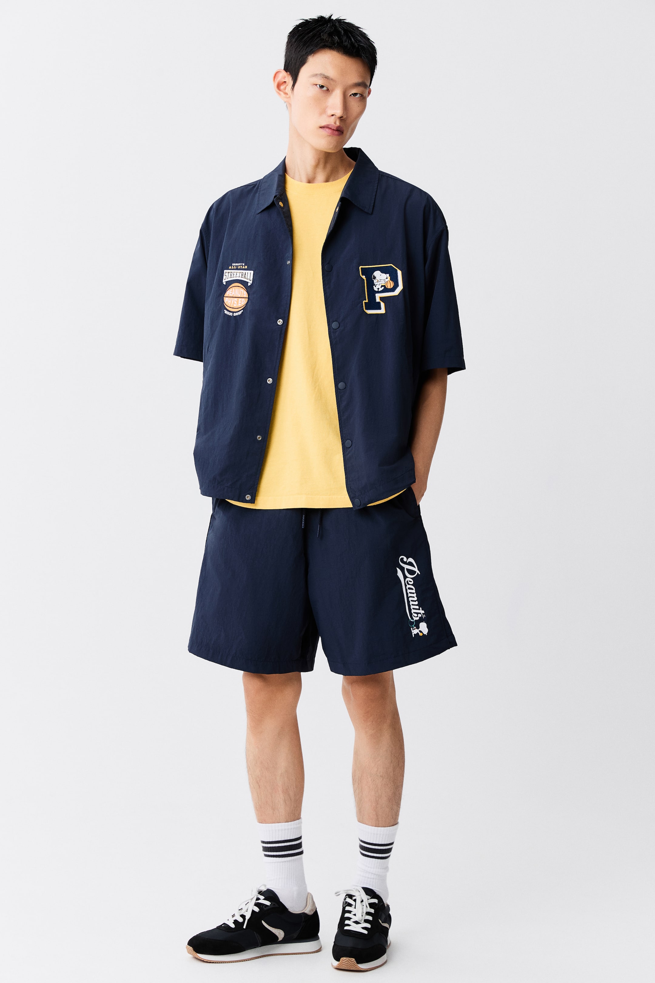 View larger image: Knee-length navy blue track shorts in crisp nylon are being worn by the model, featuring a loose fit and a white script graphic on the lower left leg. The shorts are styled with a matching navy blue short-sleeve shirt worn open over a yellow T-shirt.
