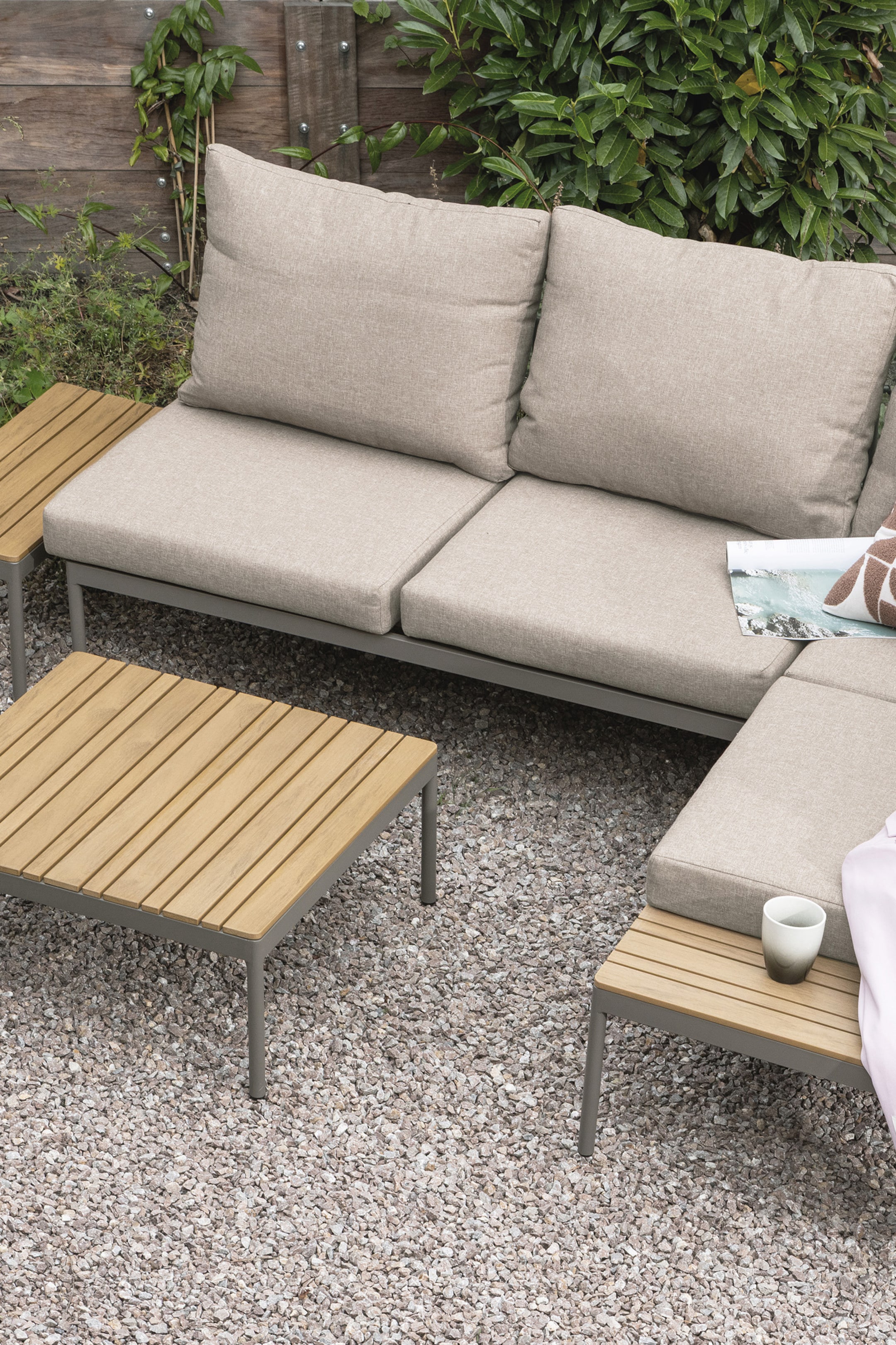 Agrandir l'image: Un ensemble de canapé modulaire d'extérieur présente une configuration en forme de L avec des coussins en tissu beige clair et des cadres en métal gris foncé. Des tables d'appoint en bois à lattes intégrées sont visibles à chaque extrémité du canapé, et une table basse carrée assortie est positionnée à l'avant.