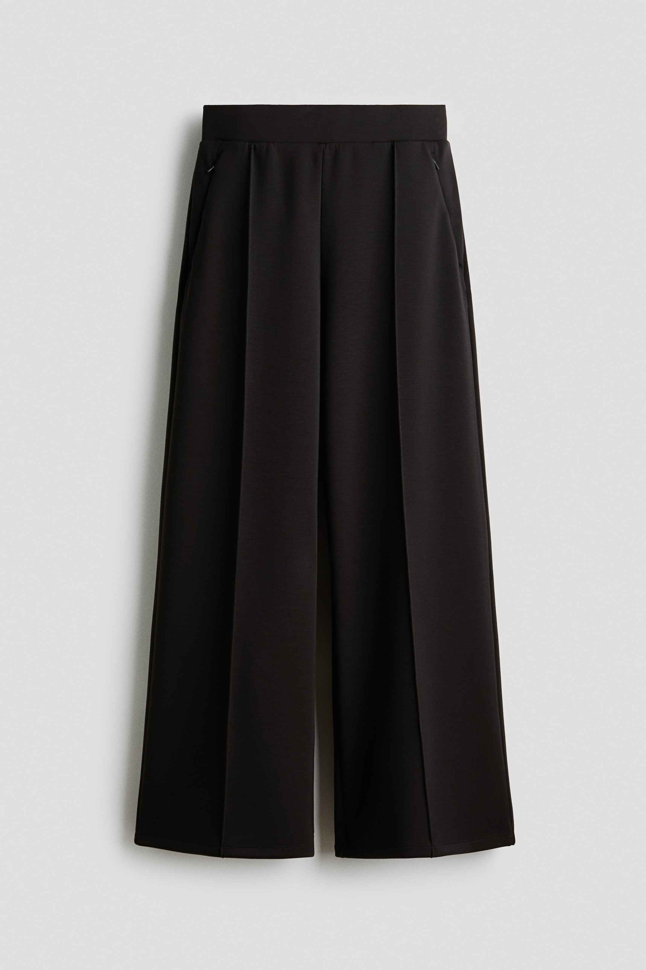 Ampliar la imagen: Pantalones negros se presentan desde el frente, con una pretina plana, piernas anchas con pliegues verticales y bolsillos laterales diagonales.