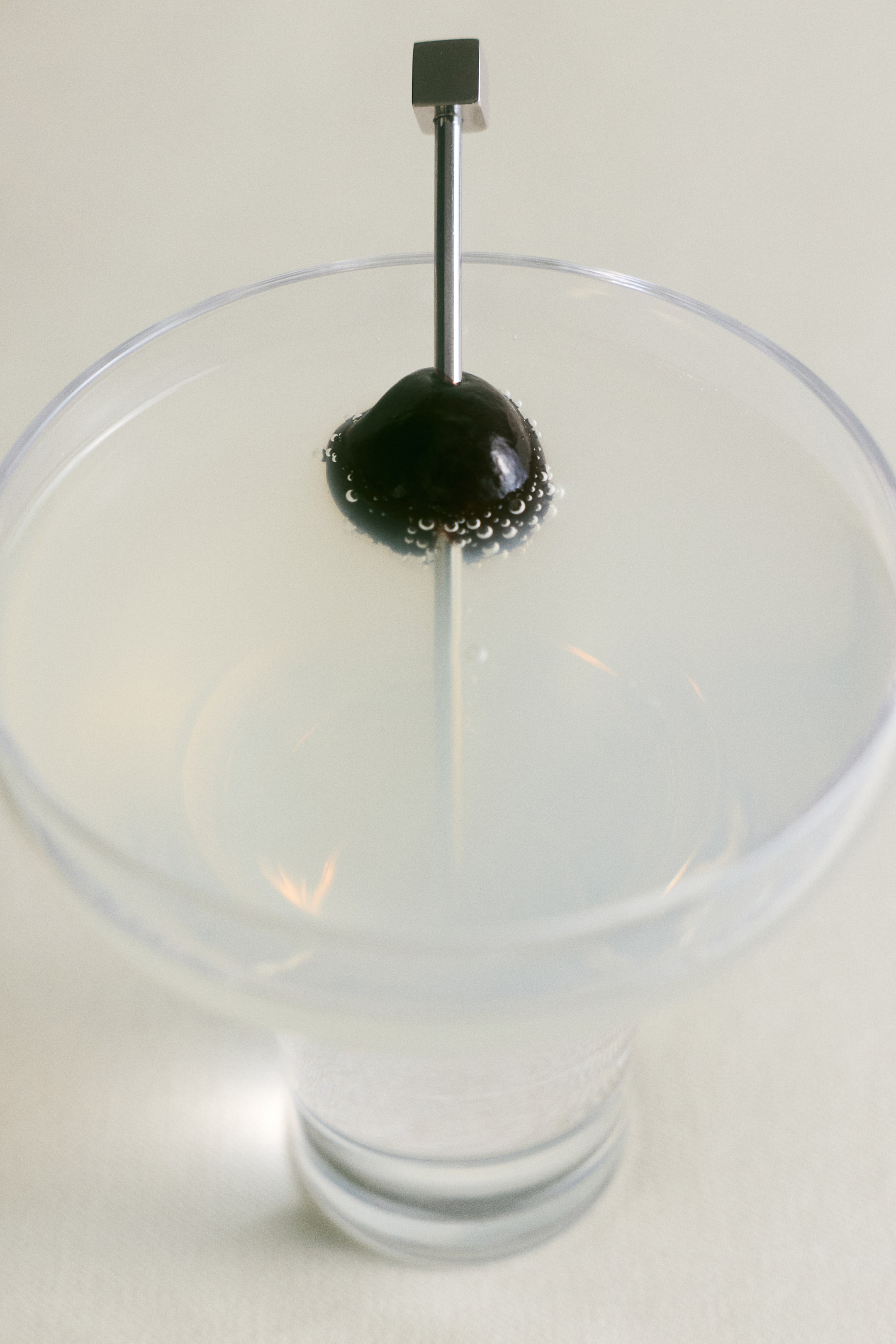 View larger image: A stainless steel cocktail pick, shown from a front-facing angle, is skewering a dark cherry garnish inside a clear glass filled with a pale liquid, highlighting its slim metallic shaft and small cube-shaped geometric decoration on top.