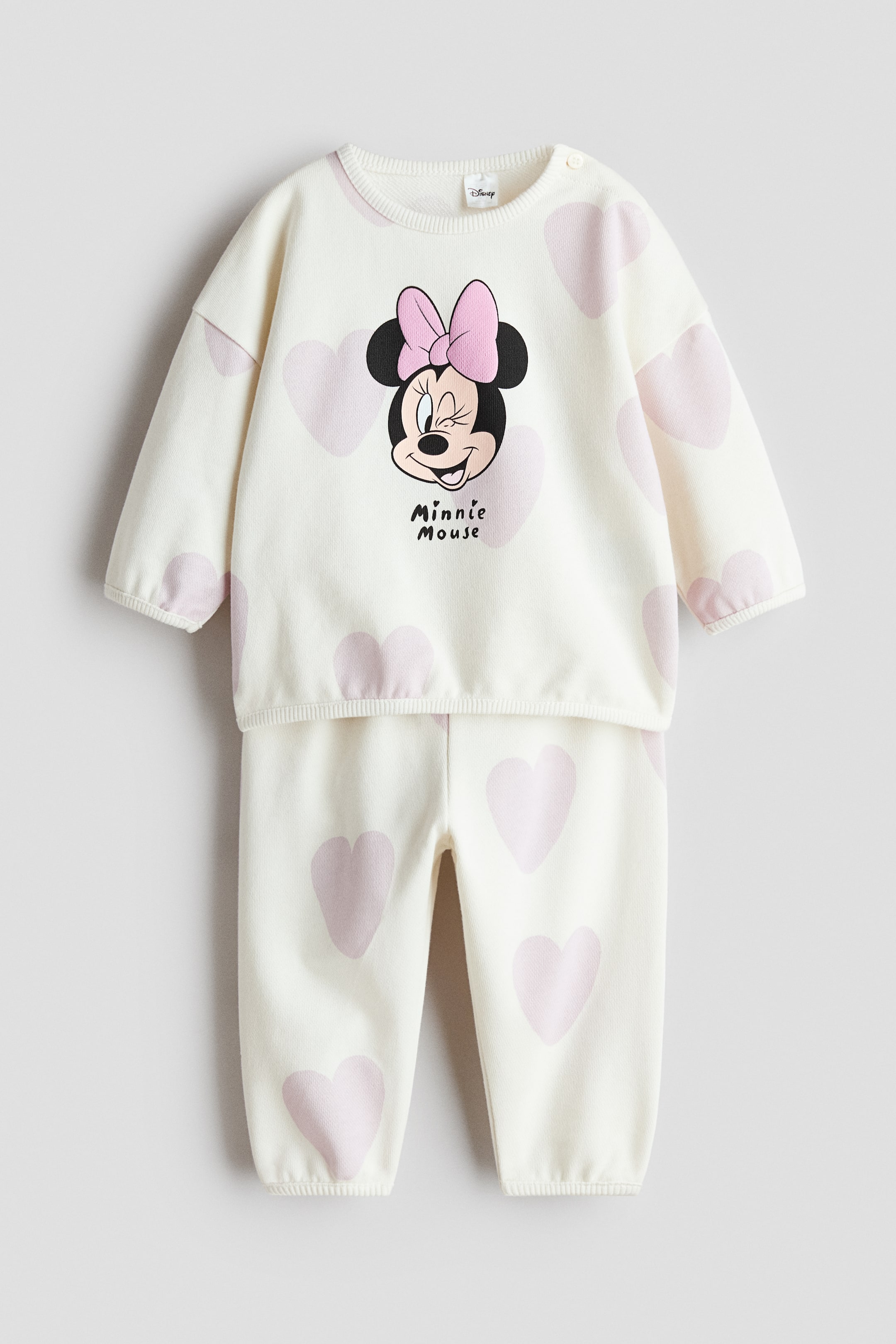 Visualizza immagine più grande: Un completo per neonati a due pezzi si presenta frontalmente, composto da una maglietta a maniche lunghe color bianco sporco e pantaloni coordinati, entrambi decorati con cuori rosa chiaro sparsi. La maglietta presenta un'immagine di Minnie Mouse con un fiocco rosa, bottoni a pressione sulla spalla e polsini a coste, mentre i pantaloni hanno caviglie elasticizzate.