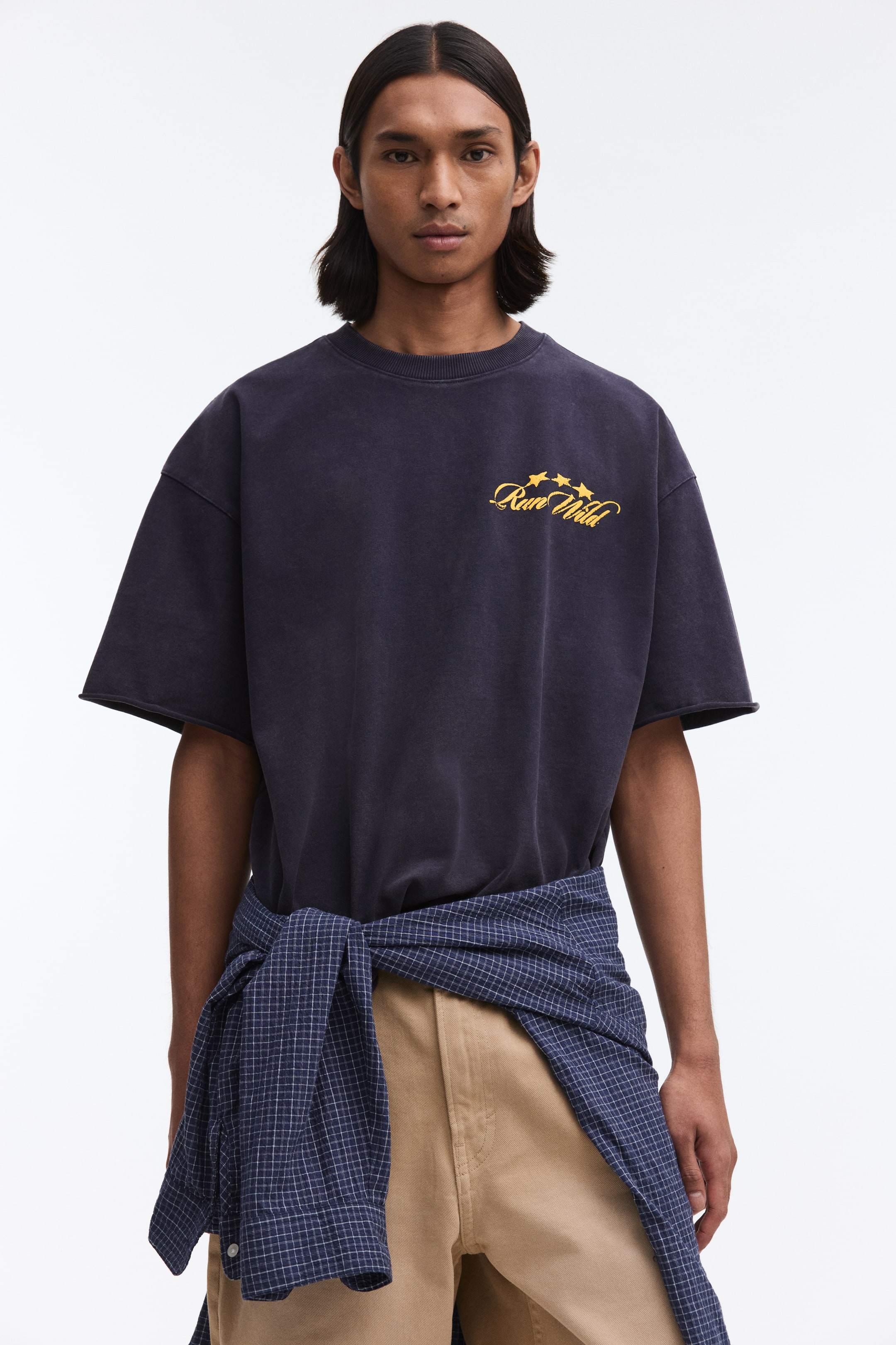 Ampliar la imagen: Una camiseta de cuello redondo azul oscuro lavado presenta un ajuste holgado y mangas cortas con bordes sin rematar, mostrando un gráfico amarillo "Run Wild" con tres estrellas en el pecho.