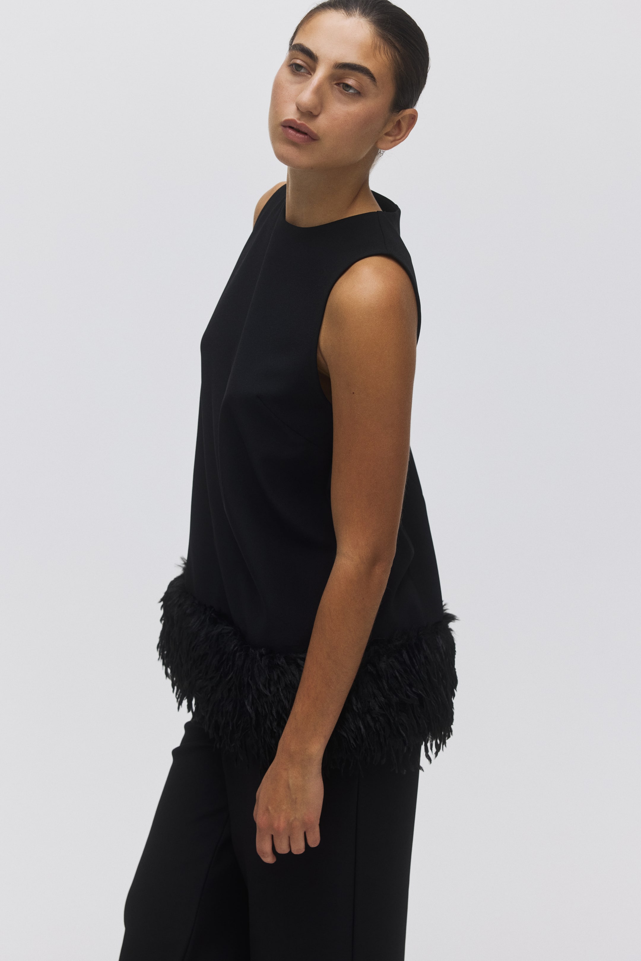 View larger image: A black sleeveless top is featuring a high neckline and a straight silhouette. It is being presented from a slight side angle, with its hemline being adorned with black feathers.