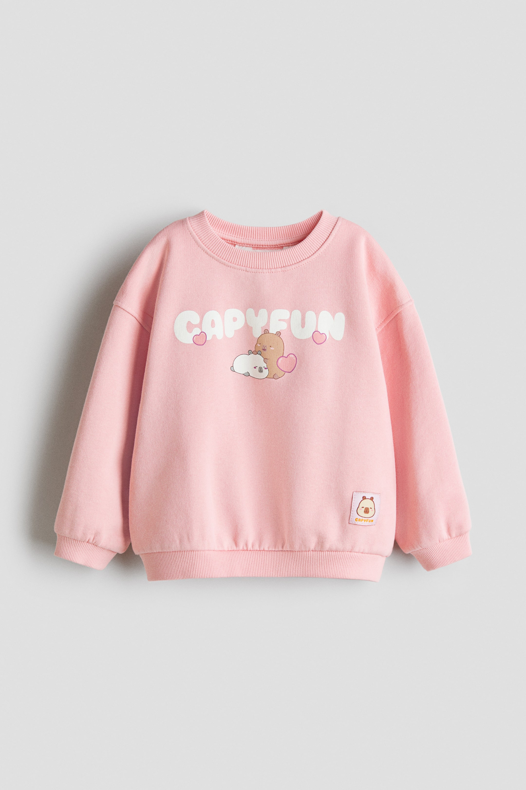 View larger image: A pink top in lightweight sweatshirt fabric is showcased at the FRONT, featuring a round, rib-trimmed neckline, dropped shoulders, and long sleeves. It boasts a large "CAPYFUN" motif with two cartoon capybaras and pink hearts, along with a small "CAPYFUN" patch with a capybara head on the lower right, complemented by ribbed cuffs and hem.