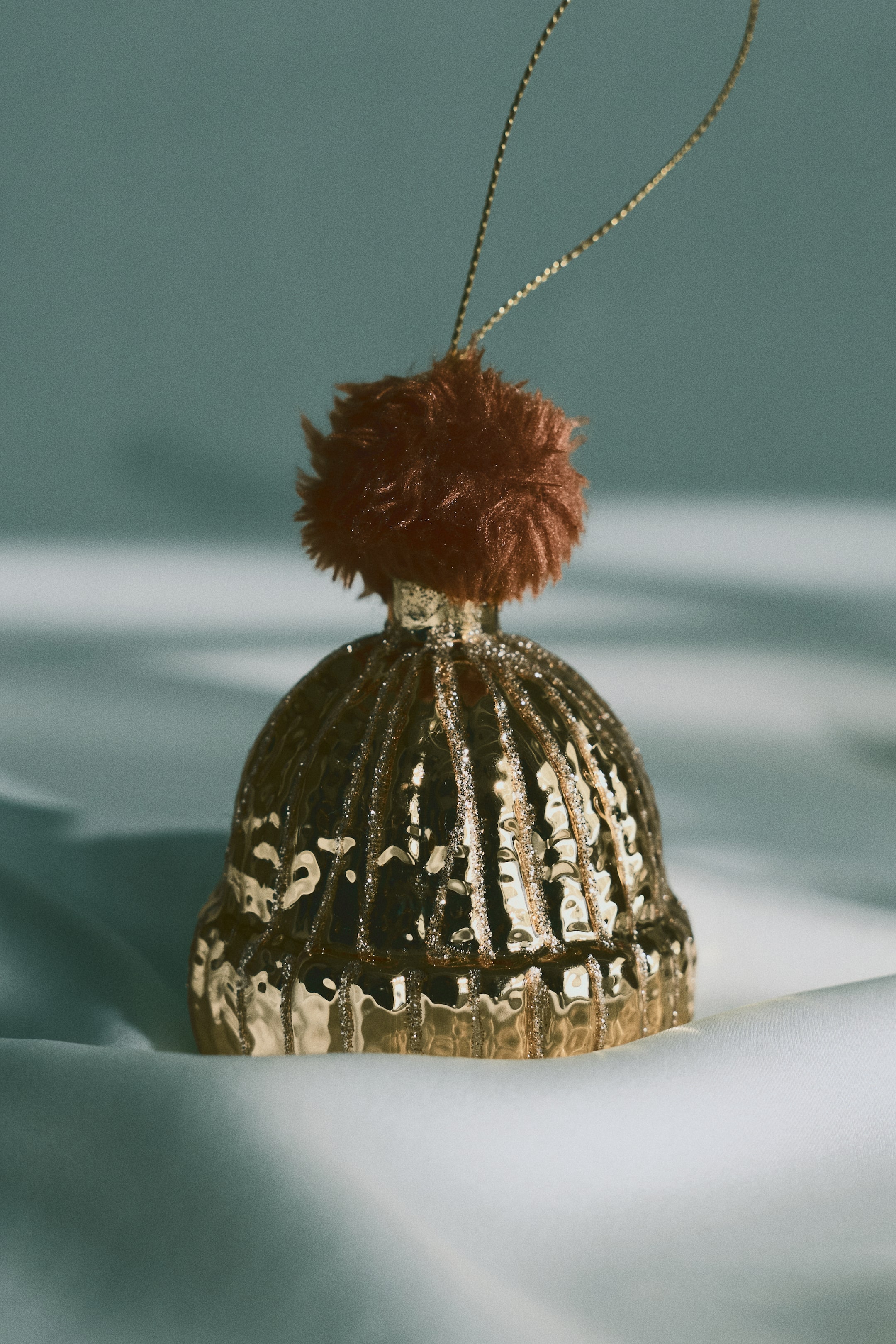 Agrandir l'image: Une décoration de Noël en verre en forme de bonnet est dressée verticalement, orientée vers l’avant. Elle présente une finition dorée brillante avec des motifs côtelés verticaux et des rayures scintillantes, et est surmontée d’un pompon brun duveteux ainsi que d’une fine boucle de suspension dorée.