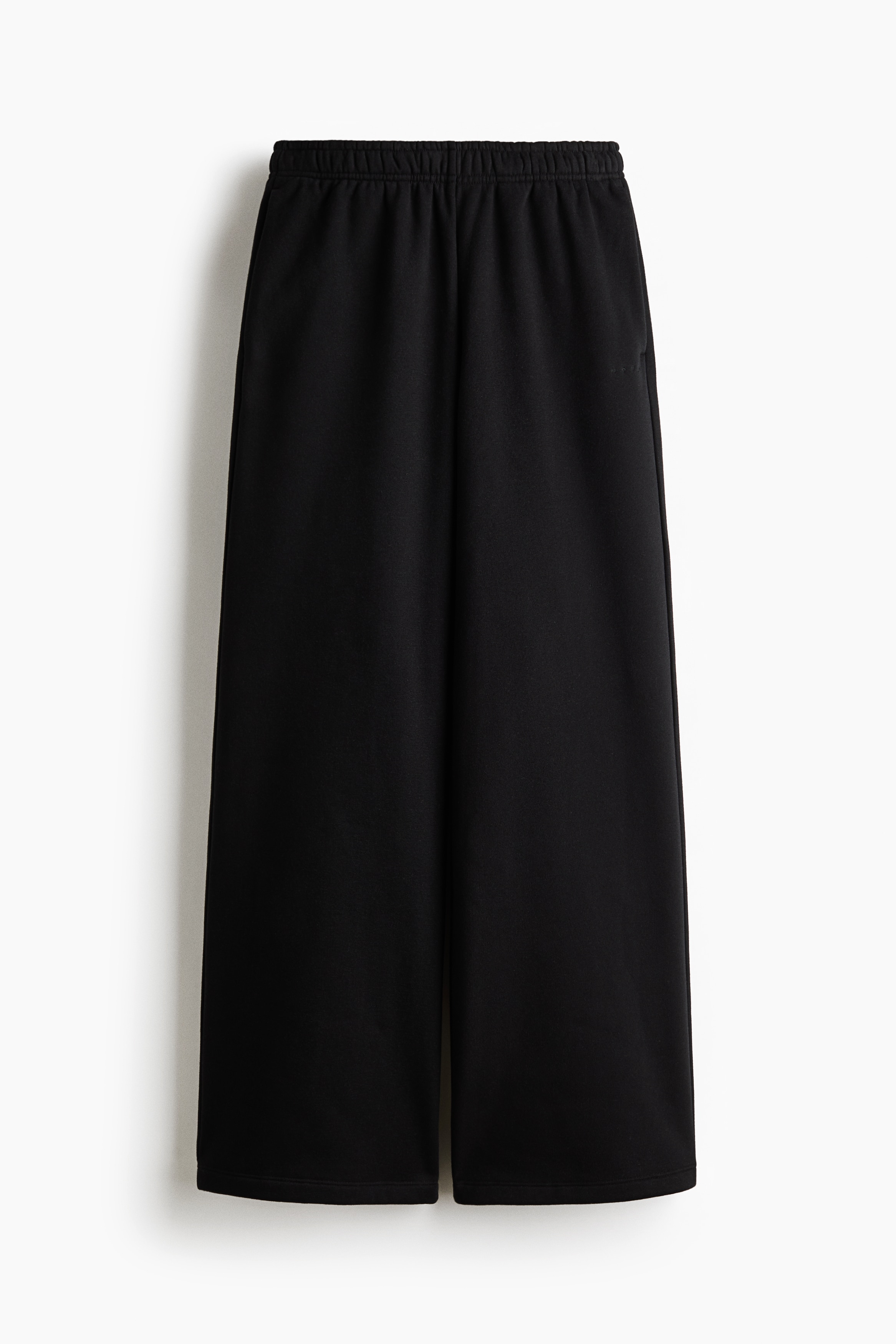 Black sports joggers featuring a wide-leg silhouette and a covered elastic waistband are being worn by the model. The model is lifting the hem of a black long-sleeve top to show the waistband of the joggers, which are styled with black trainers.