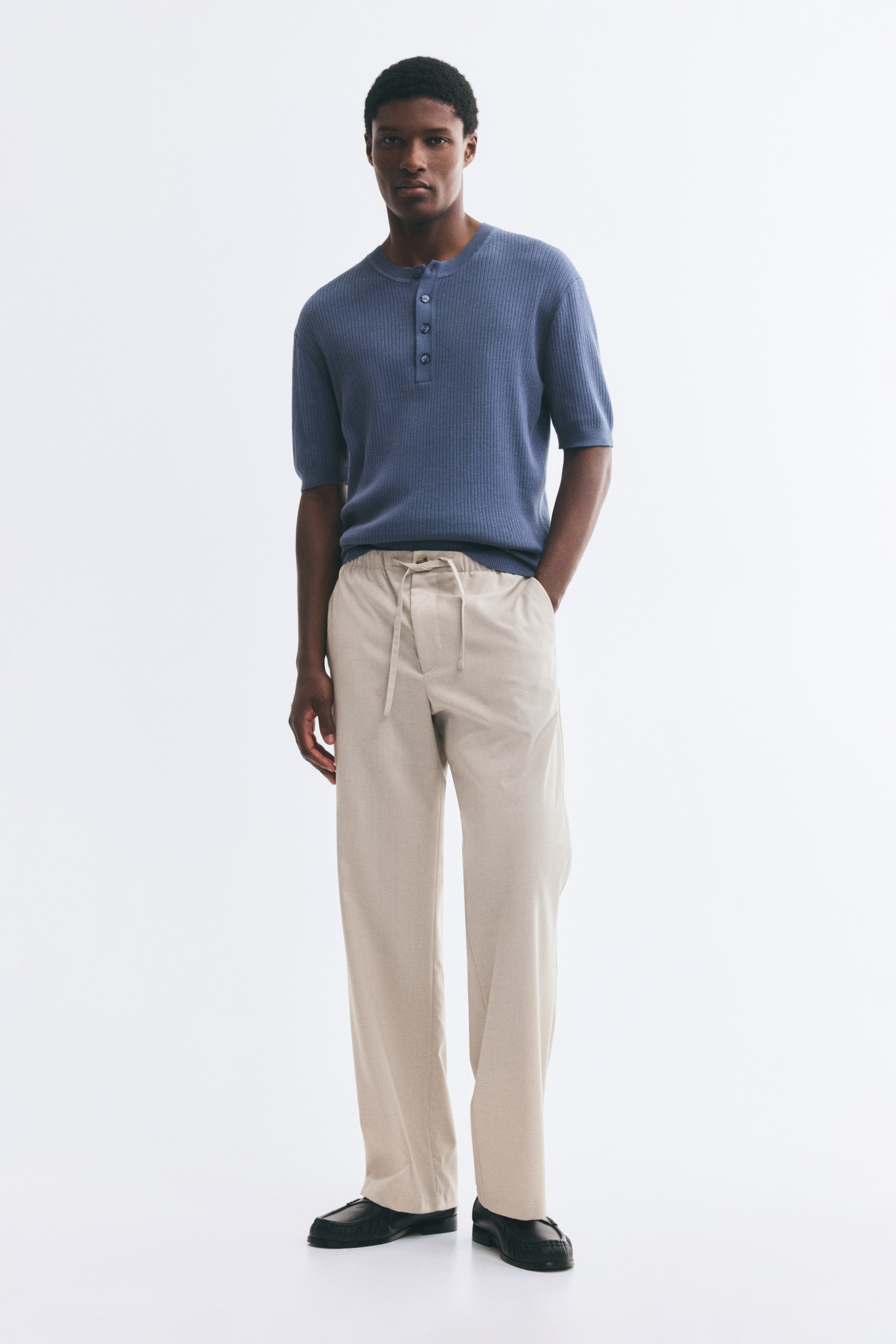 A pair of light beige trousers is shown from the front, featuring an elasticised waistband with a matching drawstring and a single button closure. The trousers fall into a relaxed, wide-leg silhouette with side seam pockets and straight hems.