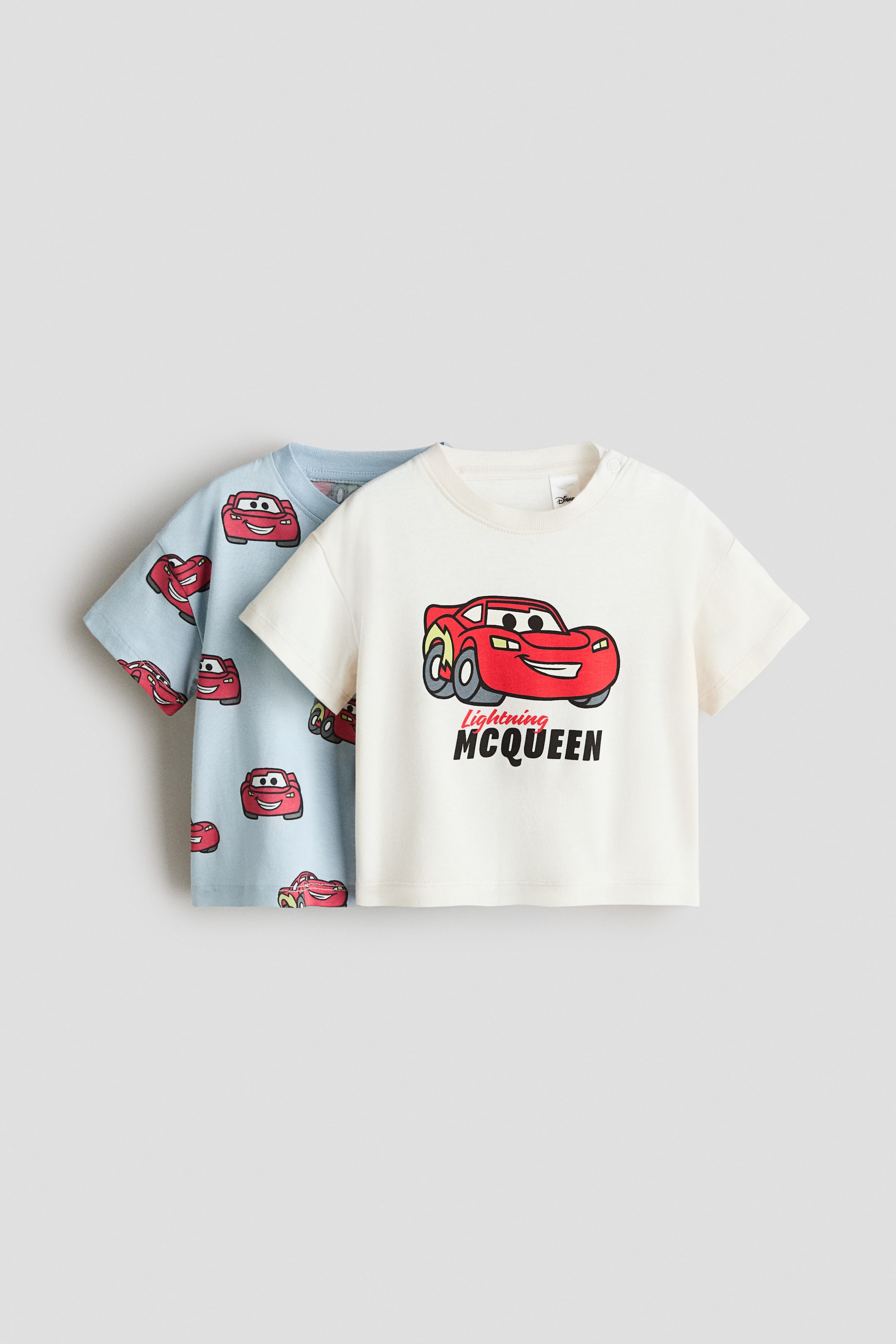 View larger image: A 2-pack of T-shirts is shown with an off-white T-shirt in front and a light blue T-shirt behind it. The off-white T-shirt features a large graphic print of the red Lightning McQueen car with the text “Lightning McQueen” below, while the light blue T-shirt displays an all-over print of smaller red Lightning McQueen cars. Both short-sleeved T-shirts feature a crew neck.