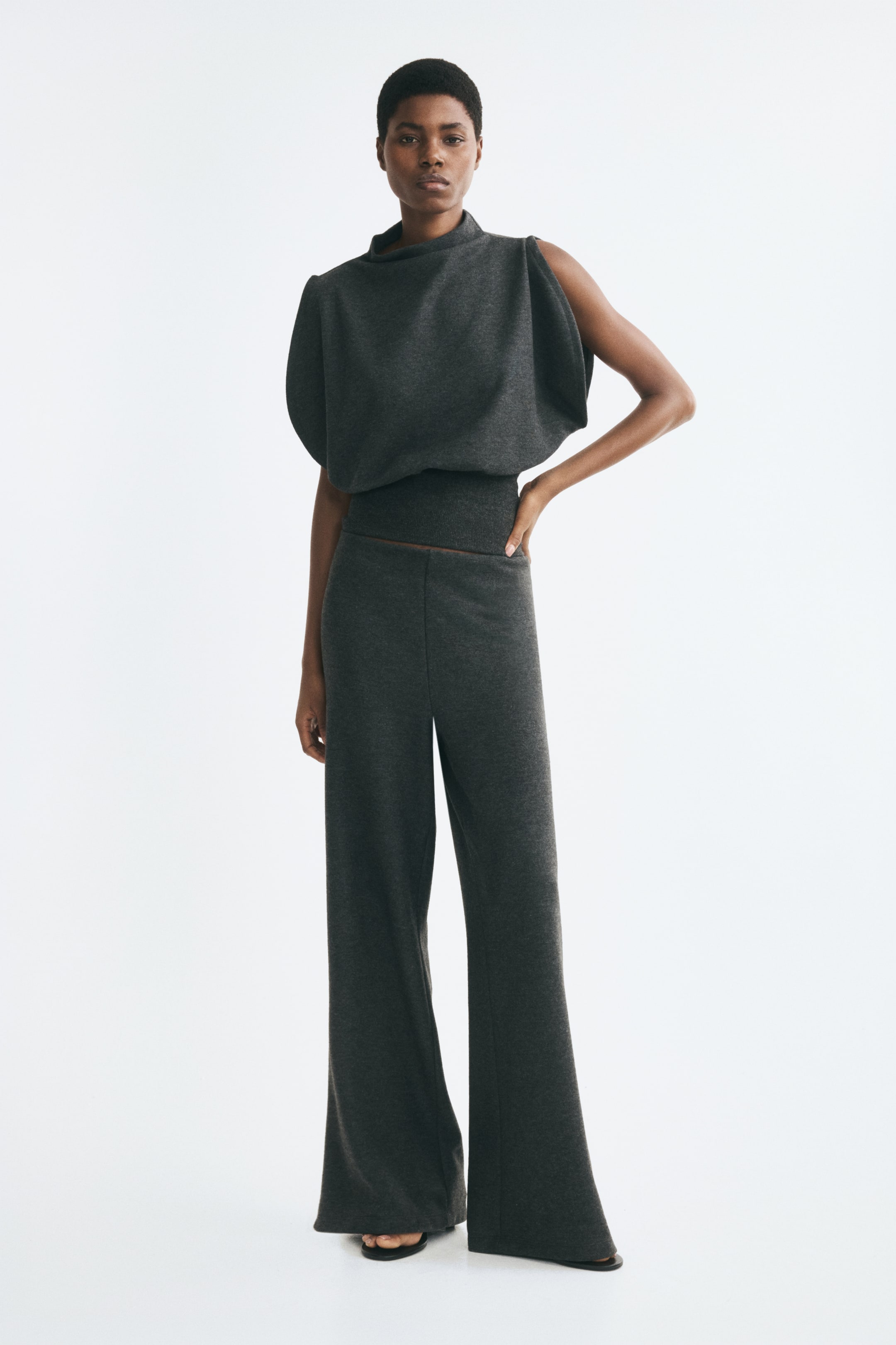 Größeres Bild anzeigen: Dunkelgraue Strickhose mit hohem, breitem elastischem Bund und geradem Bein, getragen von dem Model. Die Hose ist mit einem passenden dunkelgrauen drapierten Strickoberteil kombiniert.