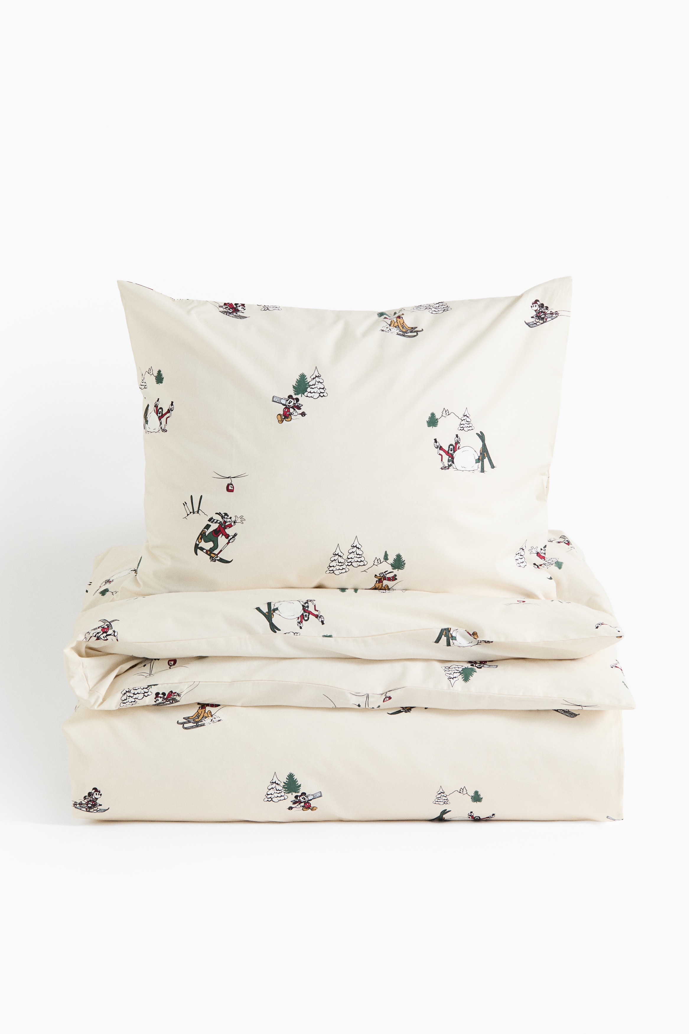 A cream-coloured cotton percale quilt cover set features an all-over winter-themed print of cartoon characters skiing, sledding, and playing in the snow. The set is shown from the front, with a matching pillowcase propped upright and the quilt cover draped softly in the foreground.