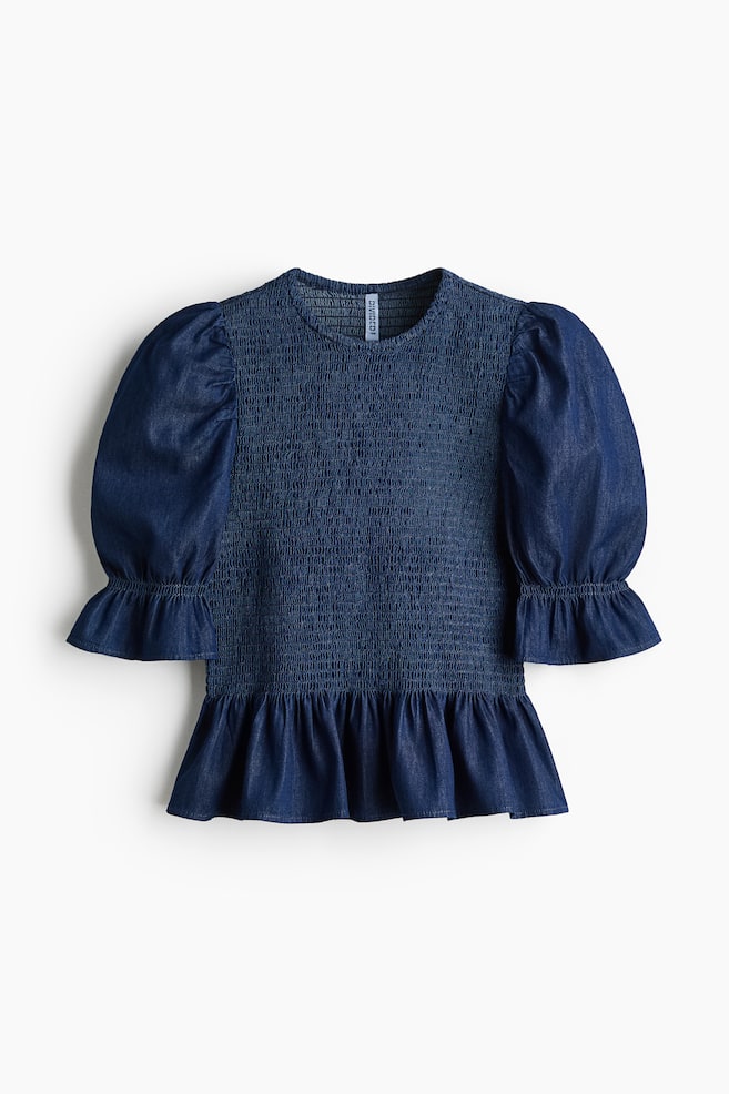 A dark blue denim smocked blouse is showcased FRONT, featuring a round neckline and short puff sleeves with narrow elastication and flared cuffs. It displays a gathered seam at the waist, leading to a flared peplum. - 1