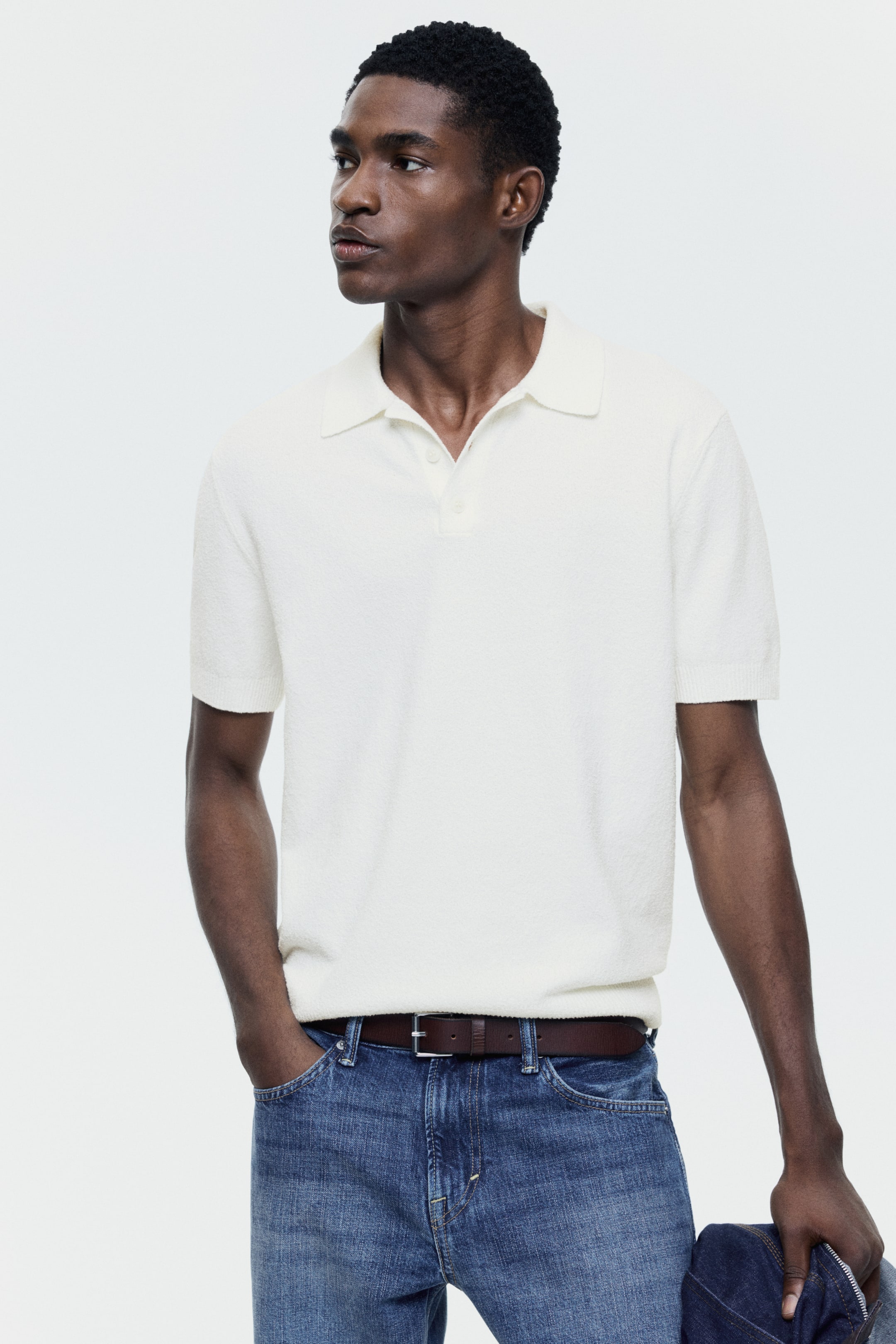Ver imagen más grande: Una camisa polo de punto texturizado en color blanco roto se presenta desde una perspectiva frontal ligeramente angulada, con un cuello clásico y una tapeta de dos botones. Presenta mangas cortas con puños acanalados, y su dobladillo está metido por dentro.
