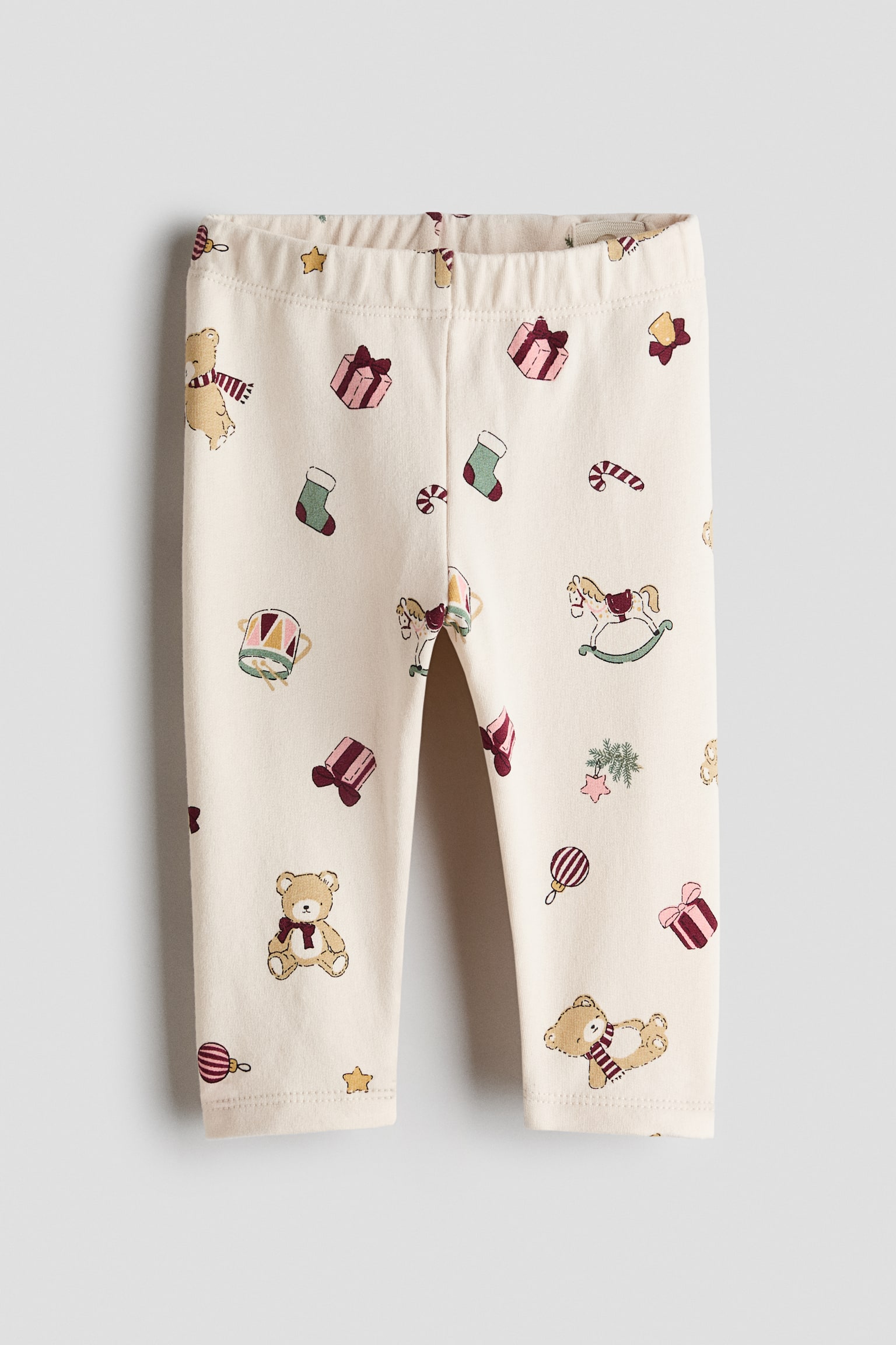 Dessa leggings i bomullsjersey i krämfärg visas framifrån och har ett allover-tryck med nallebjörnar, gunghästar, trummor och inslagna gåvor. Leggingsen har en rynkad resårlinning och raka benslut vid anklarna.