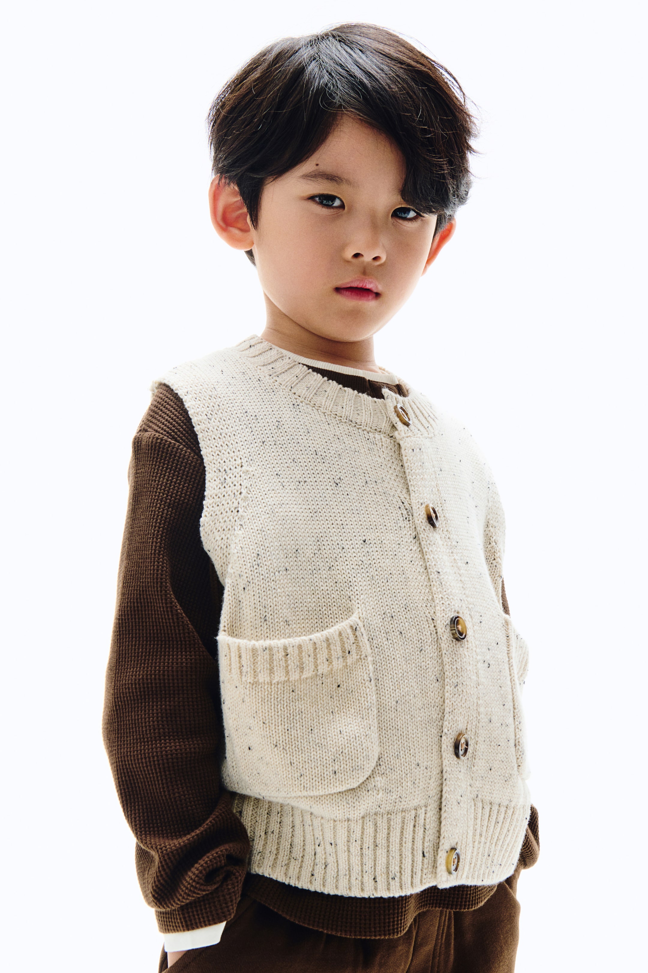 拡大画像を表示: クリーム色のニットベストにはダークな斑点があしらわれており、クルーネックとブラウンのボタンが付いた前ボタンの留め具が特徴です。それは、リブ編みの開口部が付いた2つのパッチポケットとリブ編みの裾を備えており、ブラウンの長袖シャツの上に重ねています。
