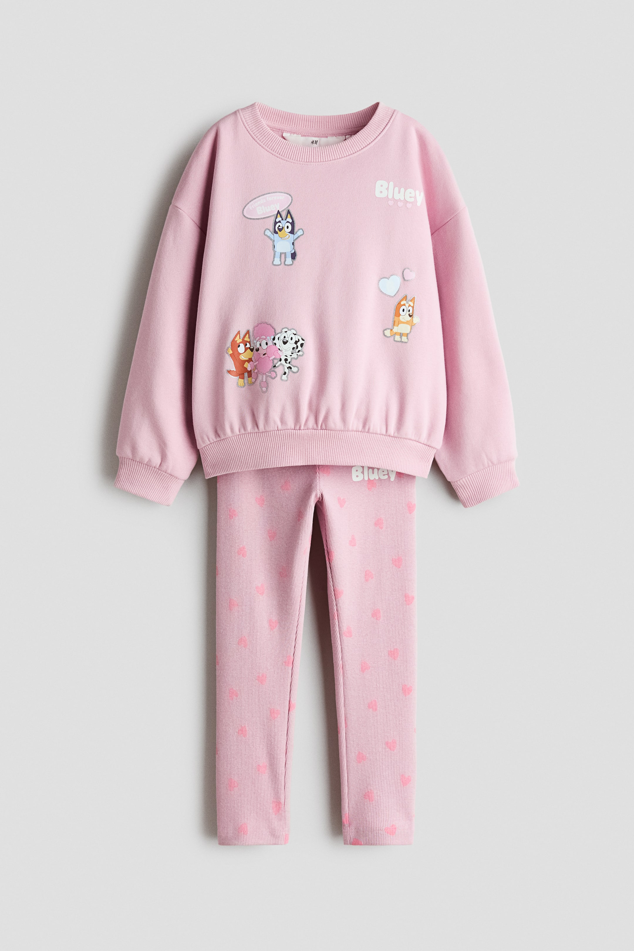 Grösseres Bild ansehen: Eine Frontansicht eines Kinderbekleidungssets zeigt einen hellrosa Pullover mit rundem Rippkragen, überschnittenen Schultern sowie Rippbündchen an Ärmeln und Saum, verziert mit aufgedruckten Bluey-Cartoonfiguren auf der Vorderseite. Die dazu passenden hellrosa Leggings zeigen ein Allover-Muster aus kleinen rosa Herzen.