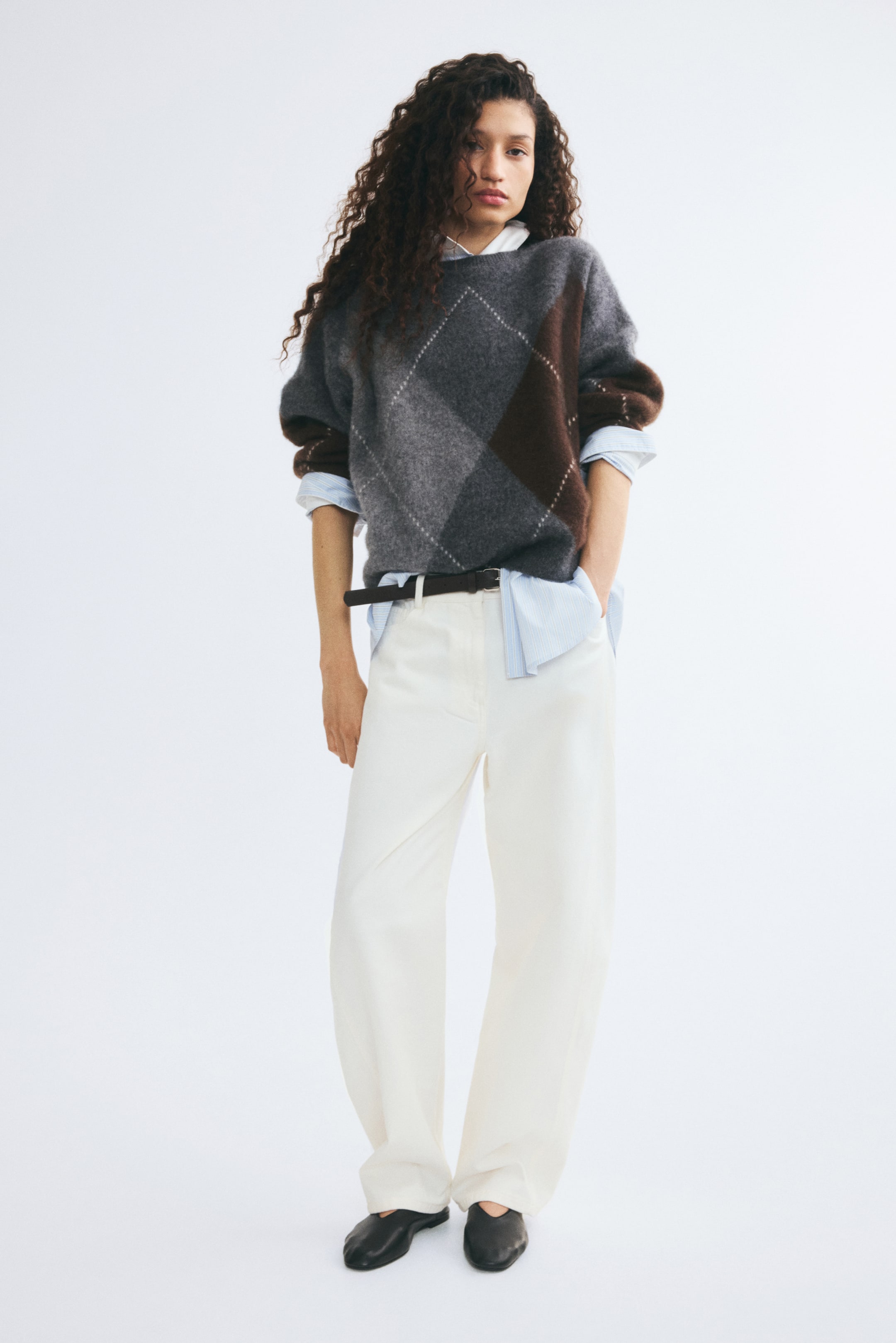 Se større billede: En oversized uldsweater i en grå og brun argyle intarsia-strik bæres af modellen, stylet over en lyseblå skjorte med krave og hvide wide-leg-bukser. Sweateren har rund hals med ribkant, sænkede skuldre samt ribstrikkede manchetter og søm.