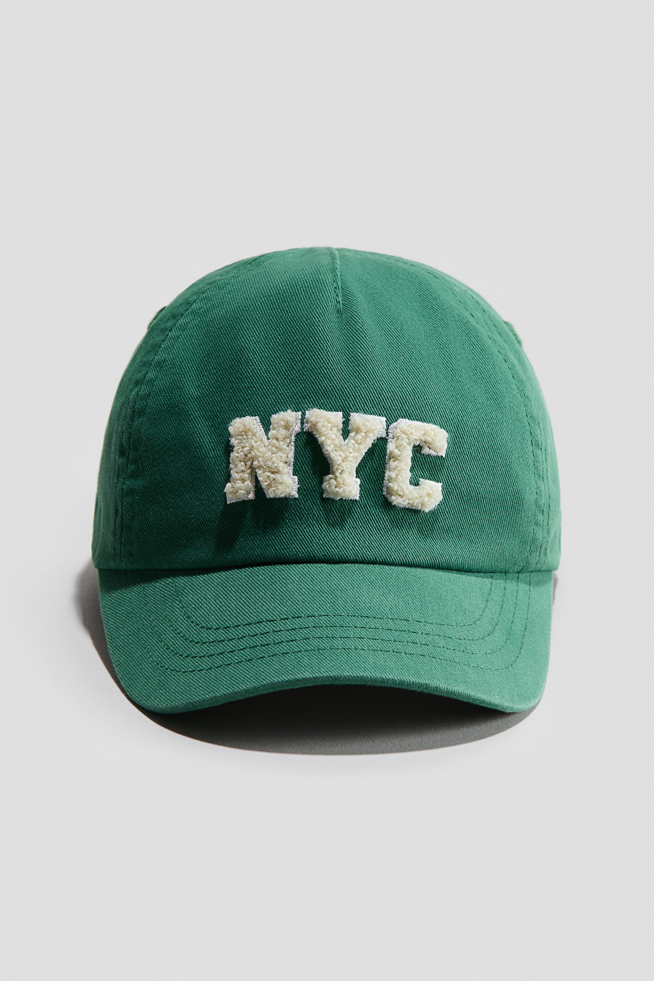 View larger image: A green cap in a cotton weave is showcasing a front view, featuring "NYC" in raised, cream-coloured chenille lettering. Its curved brim is displaying multiple stitch lines.