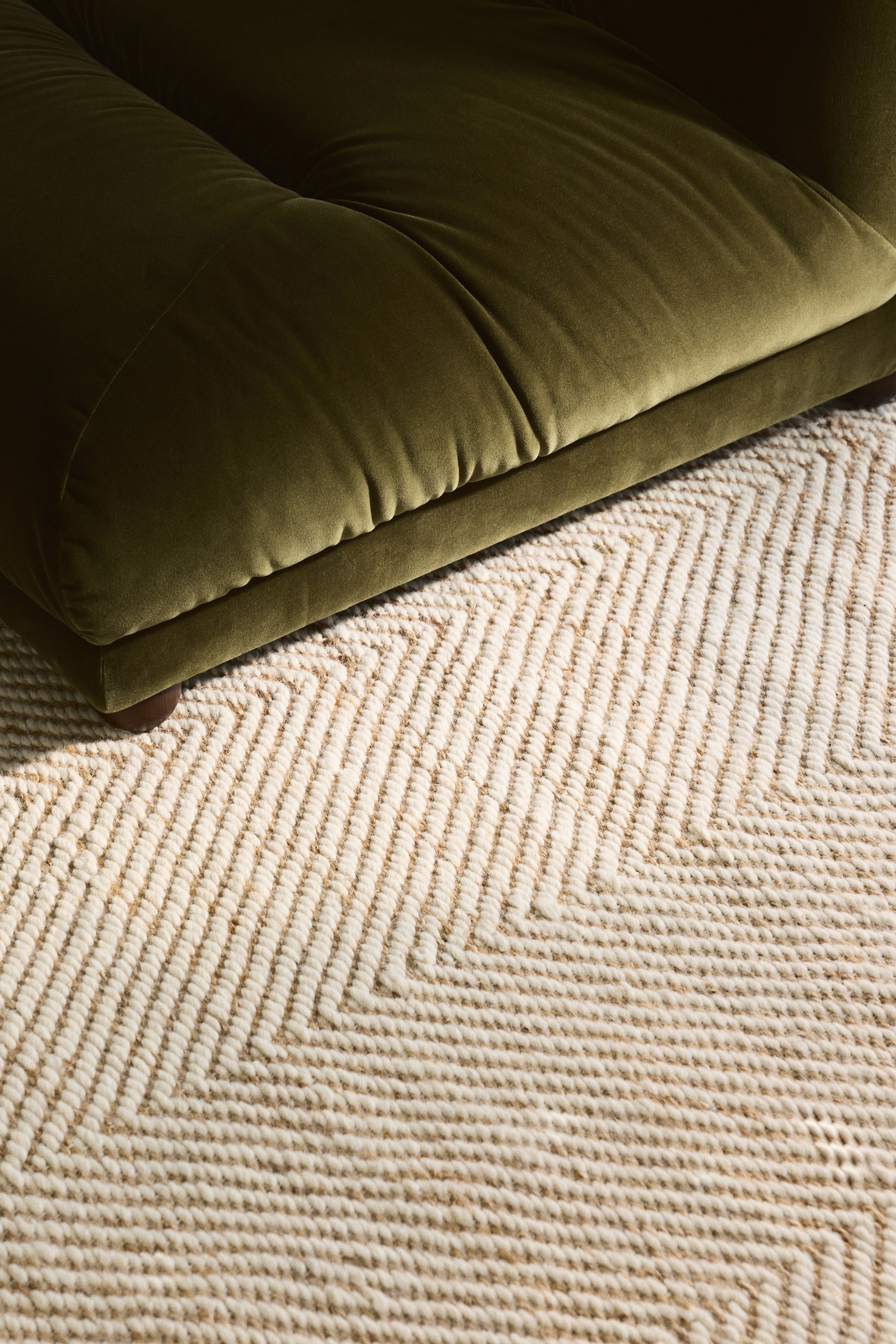Grösseres Bild ansehen: Ein cremeweißer und beigefarbener Teppich zeigt ein Chevron-Muster, dessen strukturierte Oberfläche durch dicke, gewebte Linien entsteht. Ein dunkelgrünes Sofa steht teilweise auf dem Teppich.