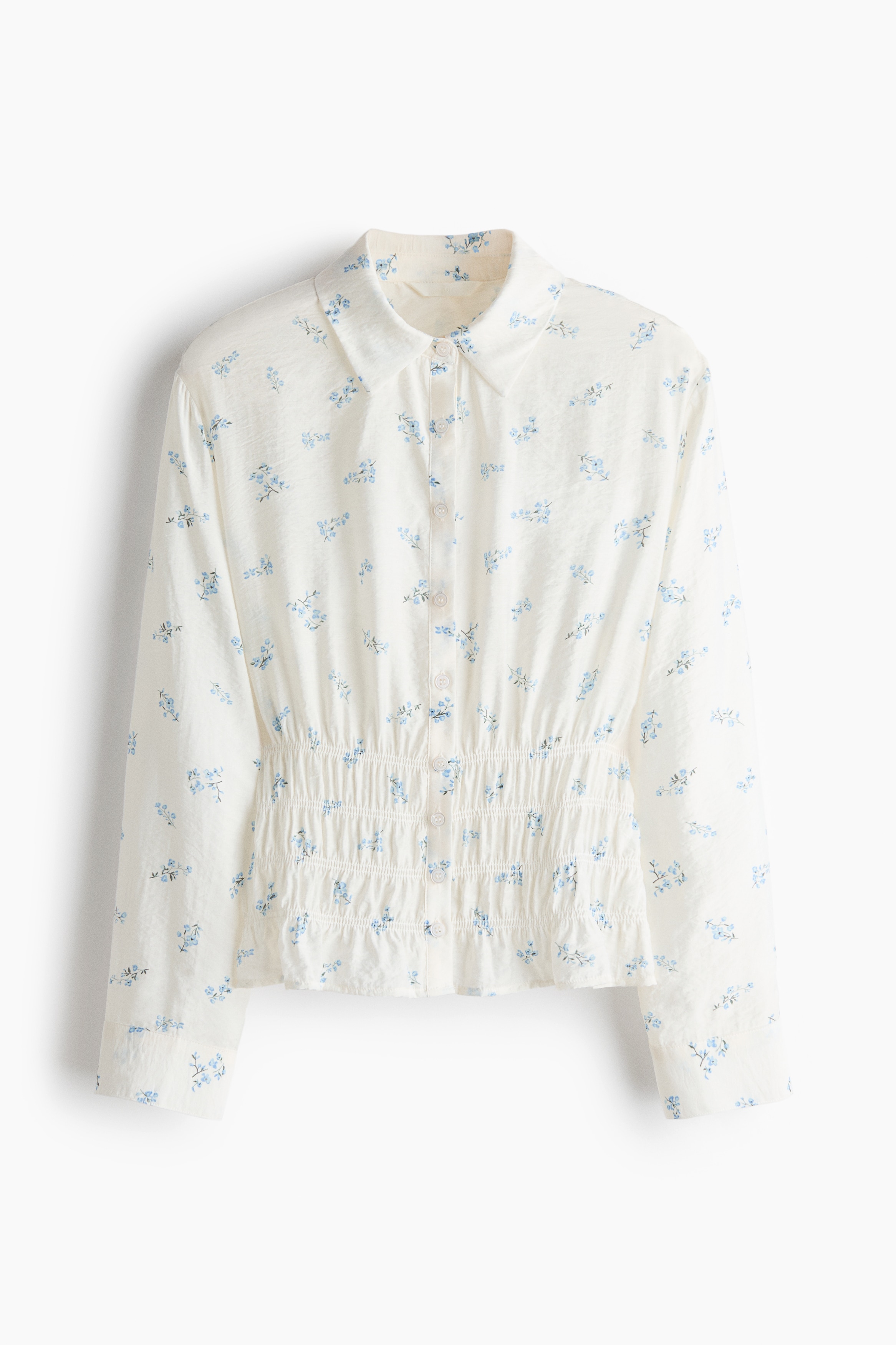 The model is wearing a white long-sleeved shirt with a blue floral print and a smocked waist. The shirt is layered under an open cream-coloured knitted vest featuring embroidered floral details, which the model is adjusting by holding its tie strings.