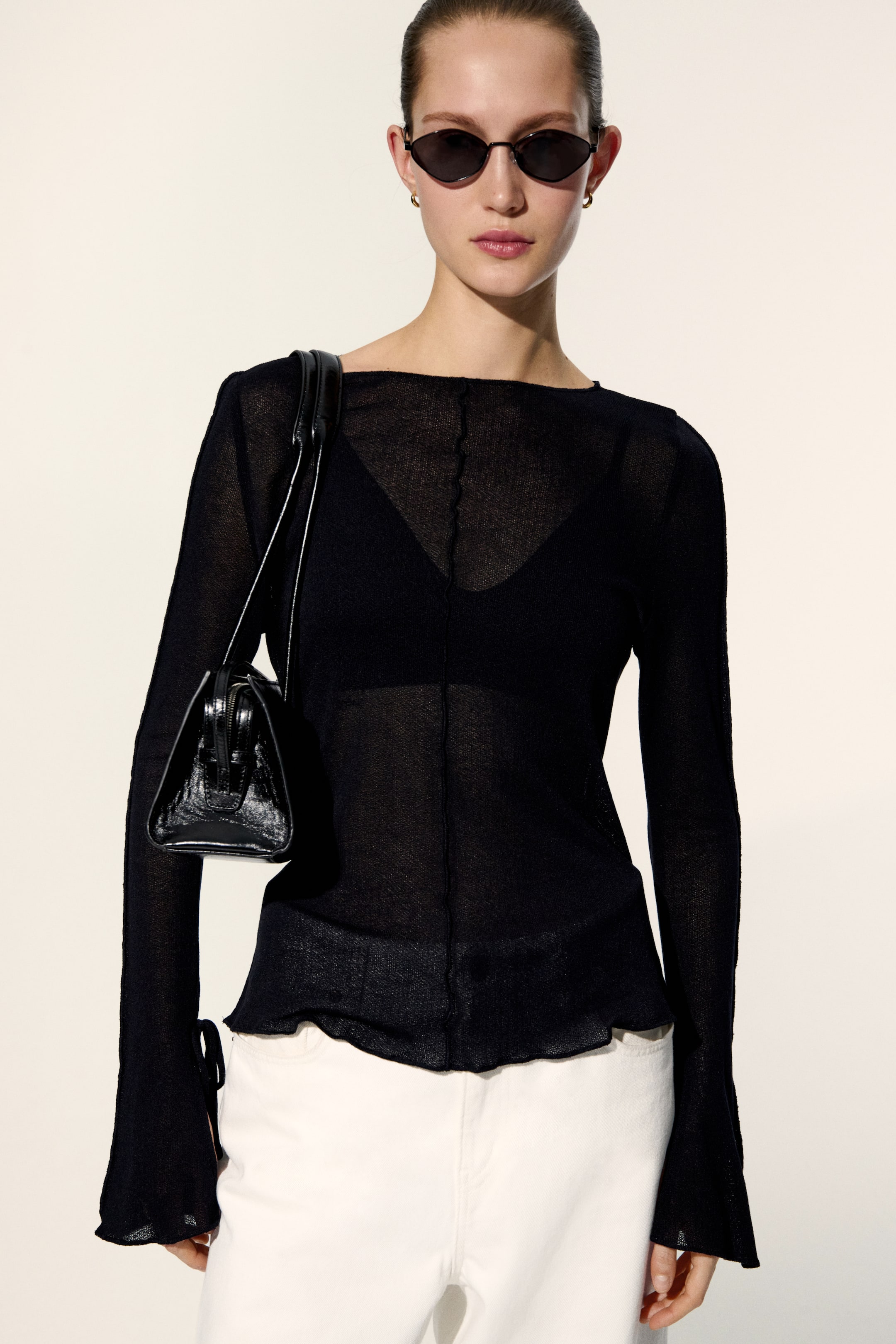 View larger image: The black sheer long-sleeve top features a wide neckline and exposed seams. Its lightweight knit creates a transparent effect, revealing a black bra underneath. The long sleeves slightly flare at the lettuce-edge cuffs, one of which showcases a tie detail, matching the lettuce-edge bottom hem.
