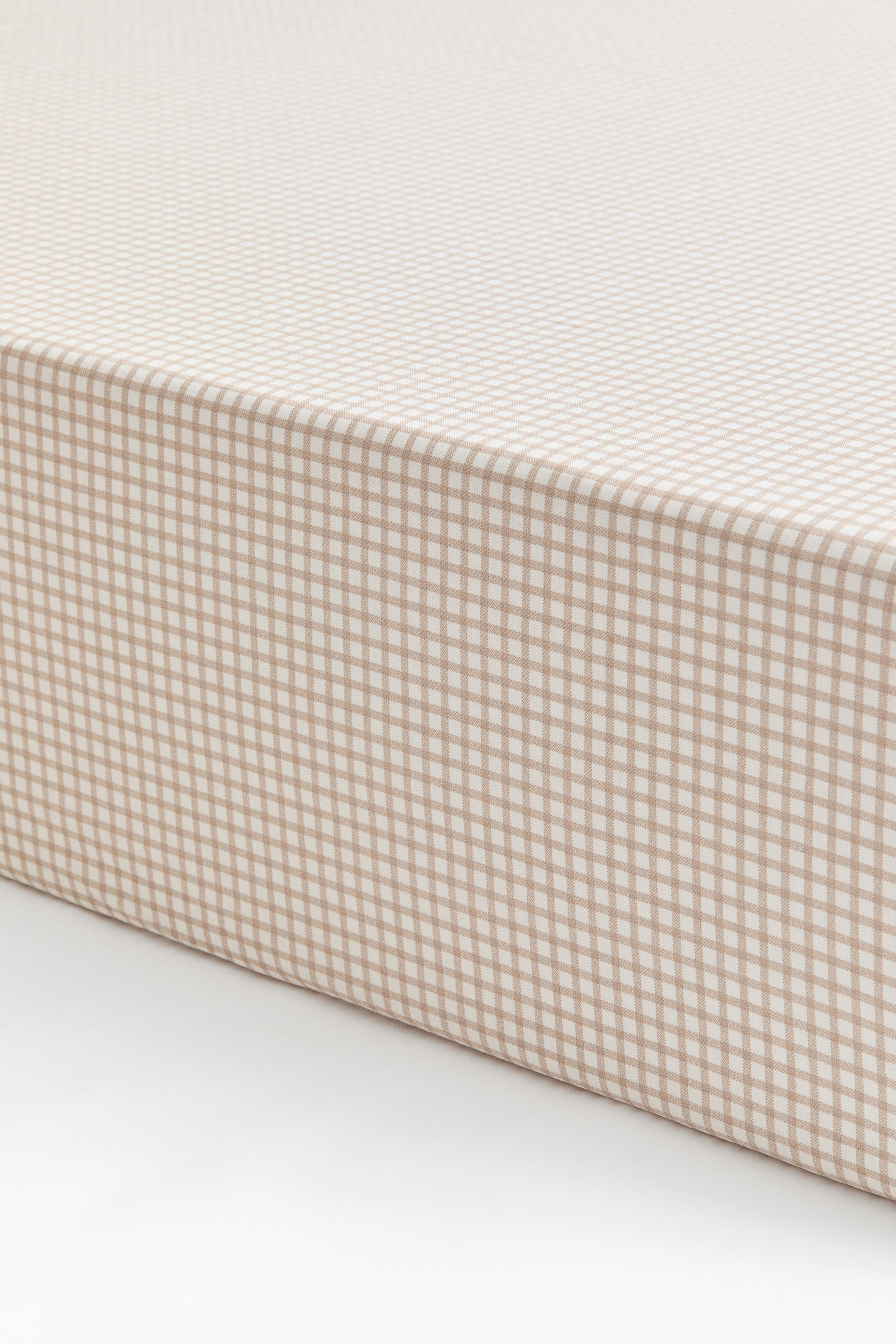 View larger image: A fitted crib sheet featuring a beige-and-white gingham check pattern is shown fitted over the corner of a mattress, displaying a smooth, snug fit along the top and side.