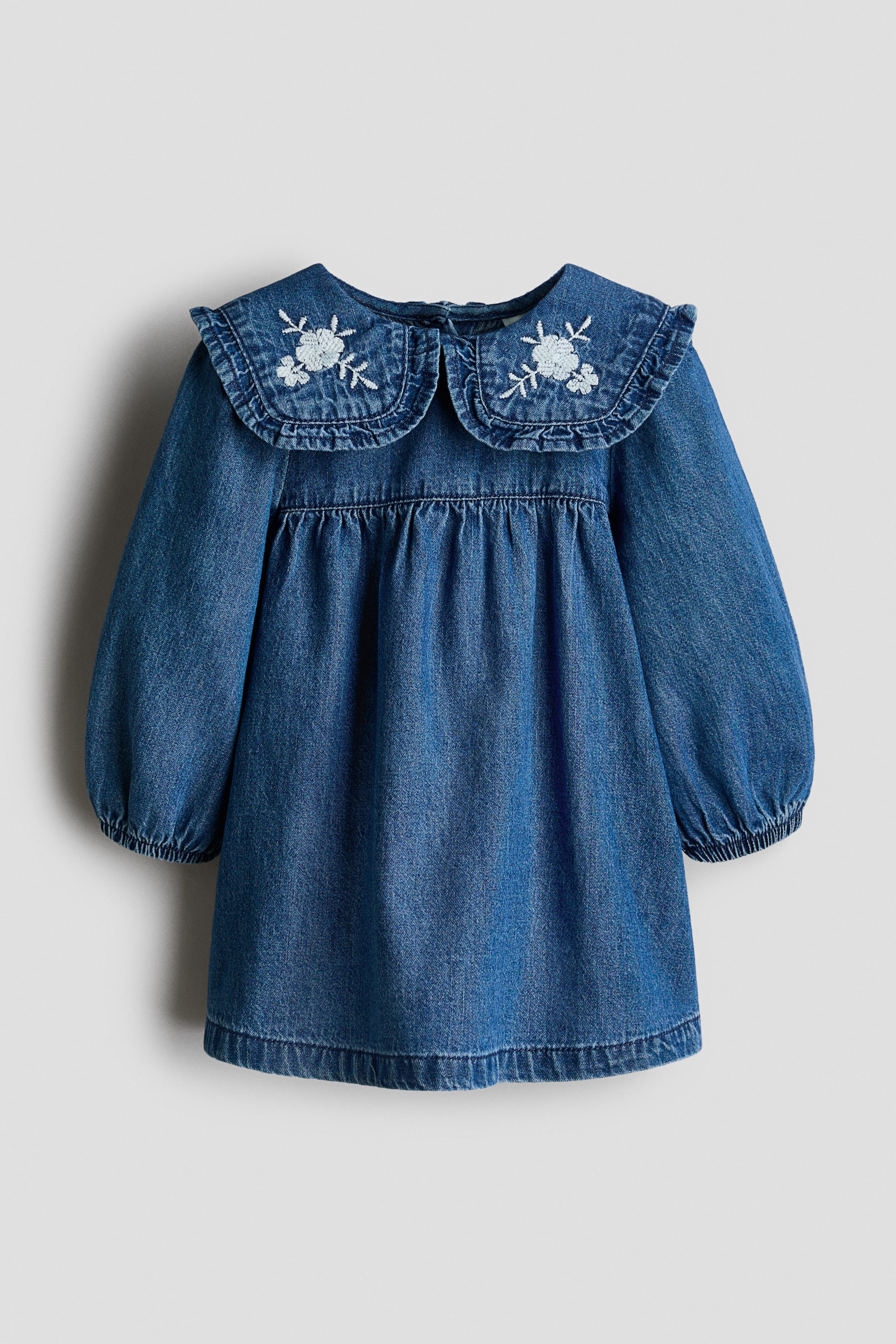 View larger image: A blue denim dress featuring a large, ruffled Peter Pan collar adorned with light blue embroidered floral designs. It has long, puffed sleeves with elasticated cuffs and a gathered waistline.