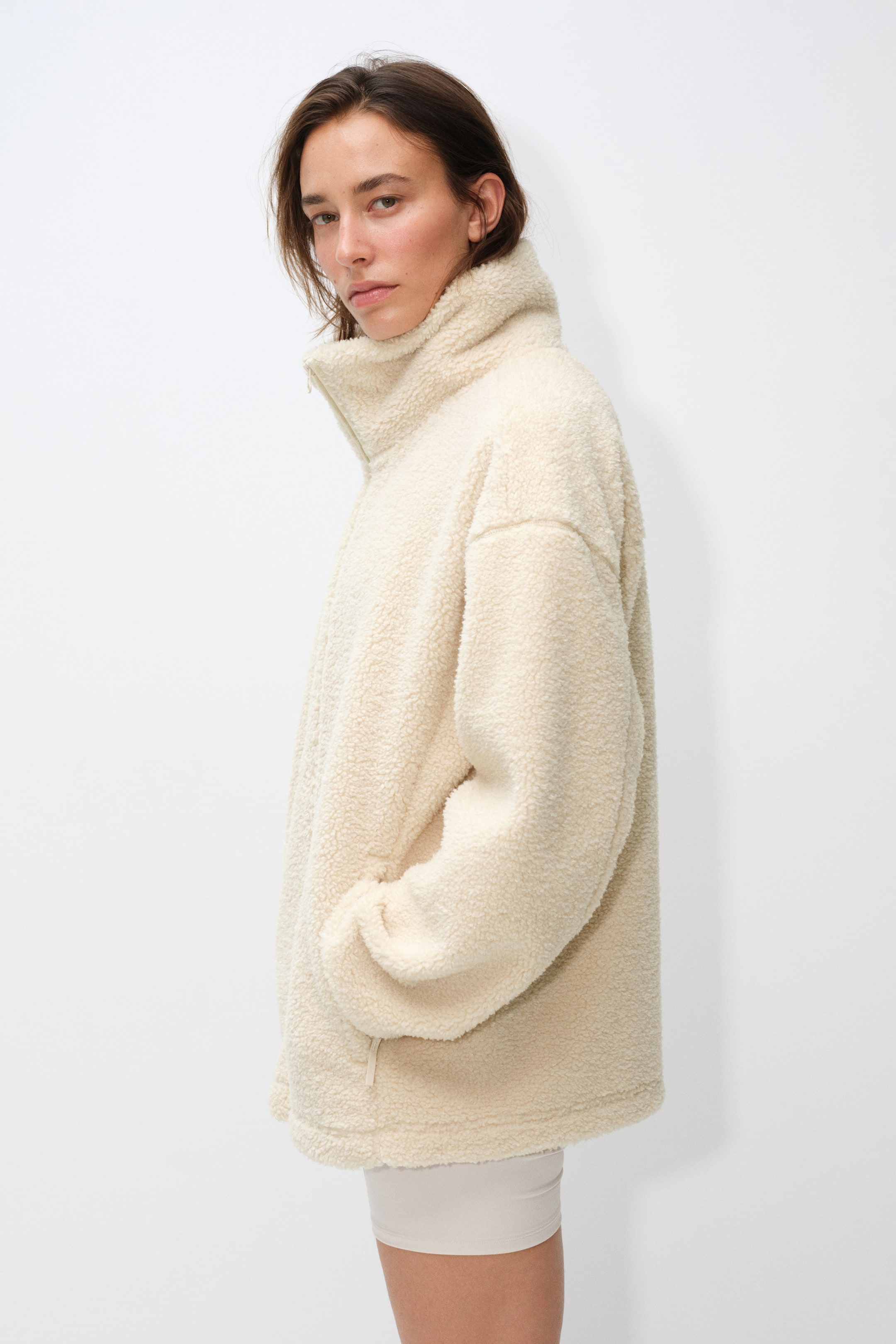 View larger image: A cream-coloured, textured fleece jacket with a high collar and front zipper is being worn by the model. The model is standing in a side-profile view with one hand tucked into the jacket’s side pocket.