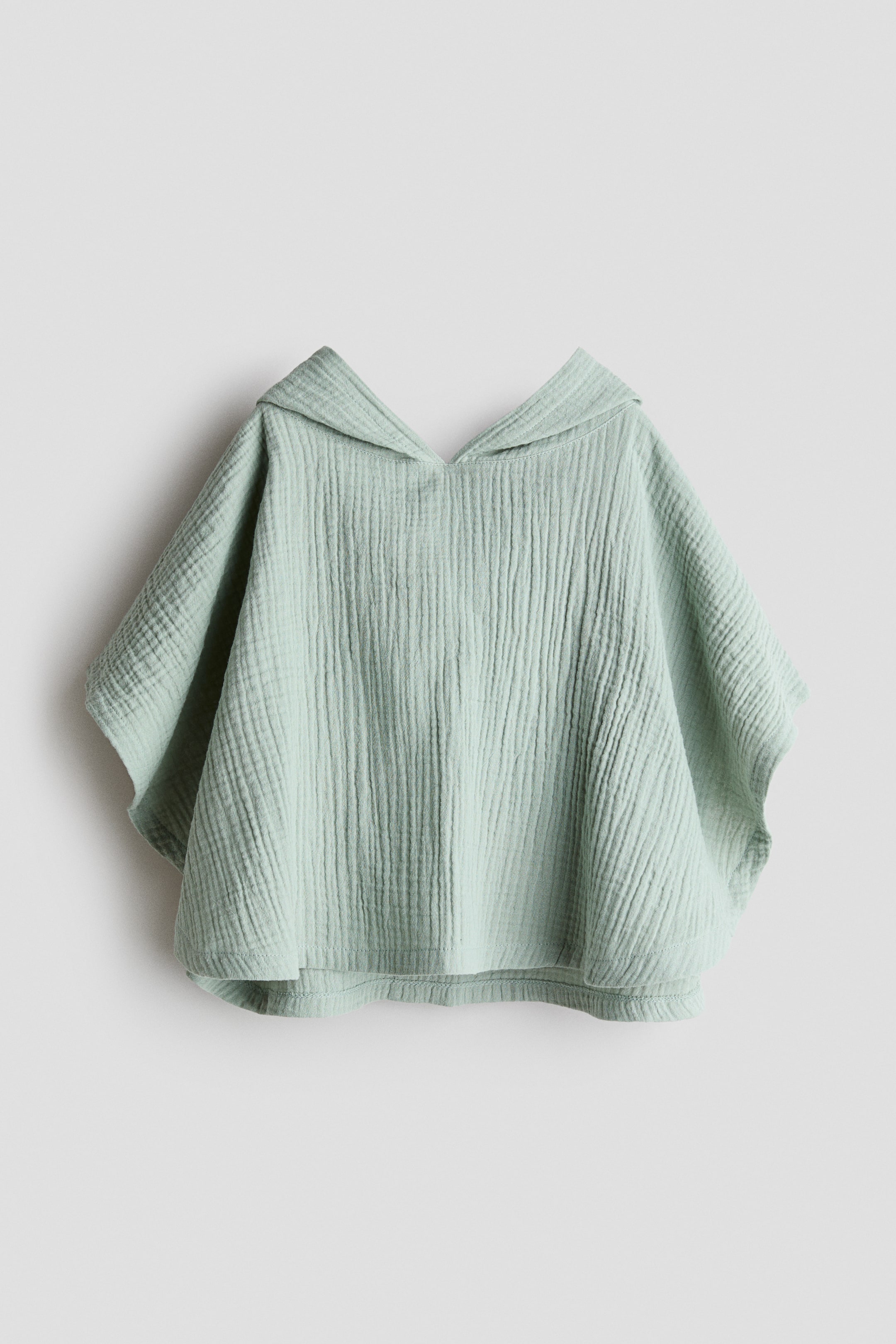Agrandir l'image: Le poncho-serviette en mousseline de coton présente une vue de face, mettant en valeur une couleur vert clair et une texture froissée caractéristique. Il est doté d'une capuche et de côtés larges et ouverts, avec un ourlet droit.