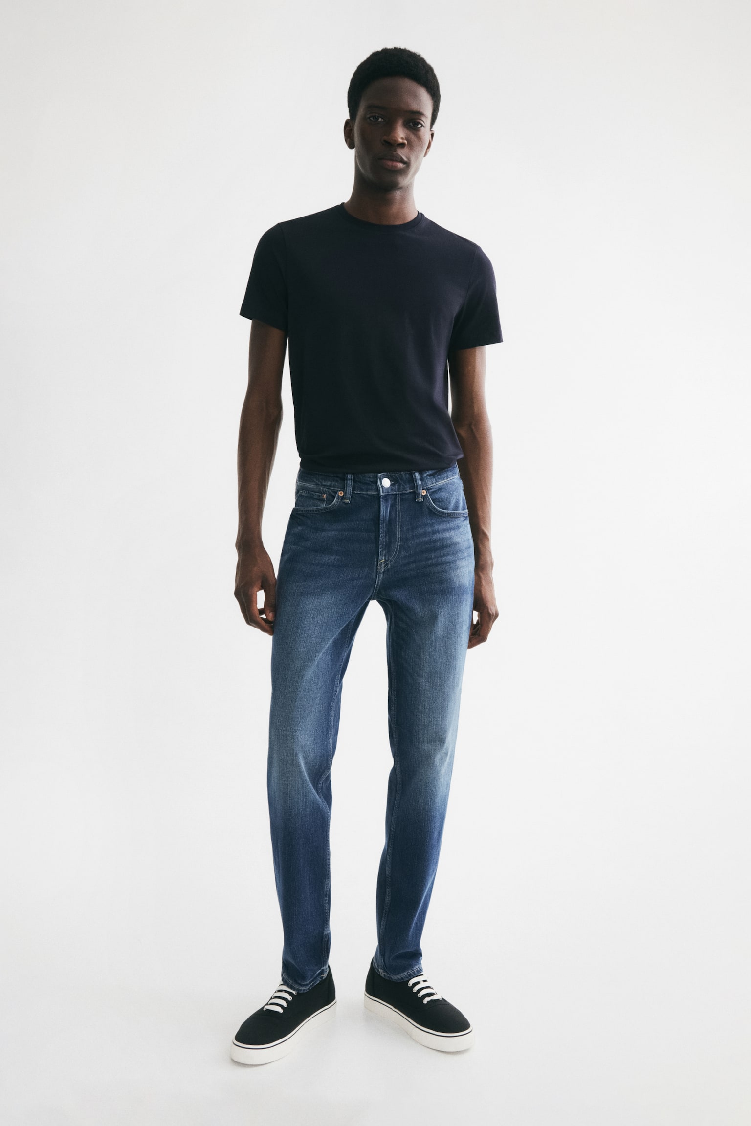 Blue denim jeans are displayed from the front, featuring a medium blue wash with lighter fading on the thighs. They are crafted with a regular waist, a button closure, two front pockets, and a coin pocket, showcasing a straight leg and a slim fit from the waist through the thigh to the hem. - 1