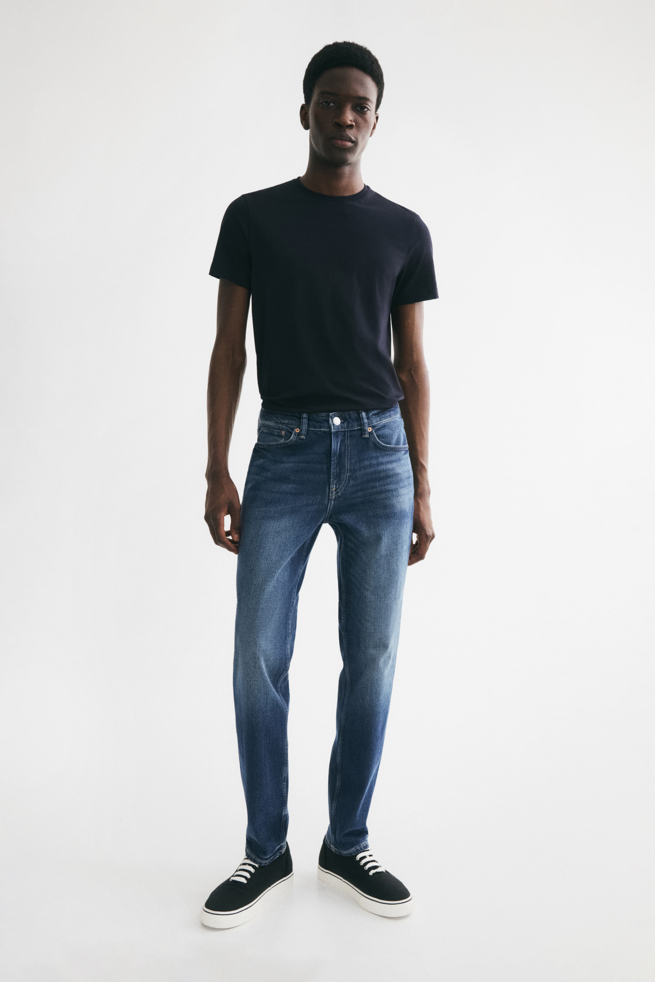 Größeres Bild anzeigen: Blaue 5-Pocket-Jeans aus Baumwoll-Denim mit leichtem Stretch, die eine reguläre Taille, einen Reißverschluss und ein gerades Bein mit schmaler Passform von der Taille über den Oberschenkel bis zum Saum aufweisen, werden vom Model getragen und mit einem dunklen, eingesteckten T-Shirt kombiniert.