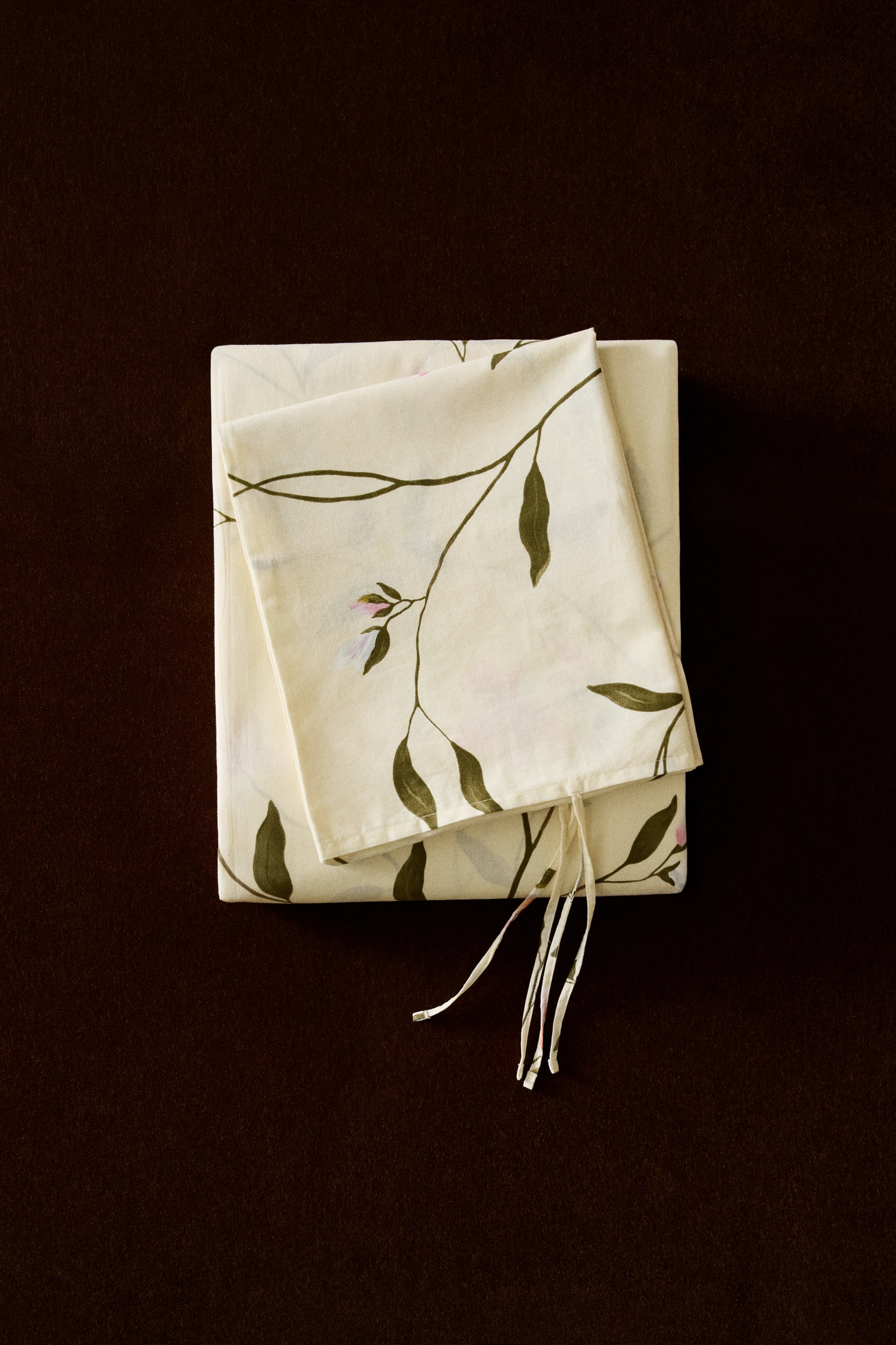 View larger image: A single duvet cover set is showcased, folded tidily, FRONT. It boasts a cream-coloured fabric adorned with a green leafy floral pattern and delicate pink blossoms. A coordinating pillowcase with ties is visible on top.