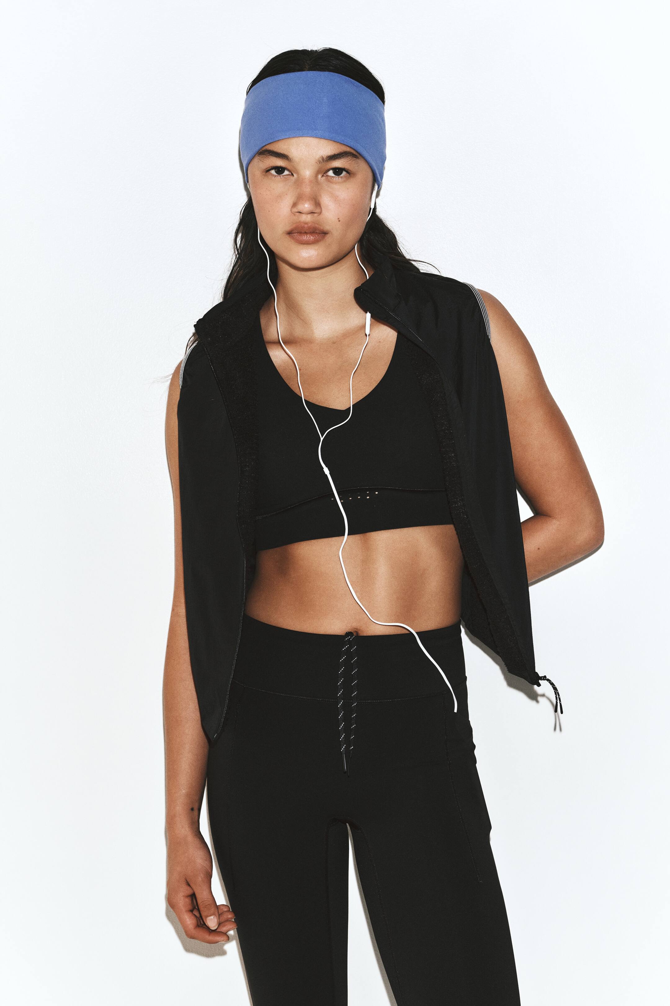 Vis større bilde: En svart treningsvest er båret åpen av modellen, lagt over en matchende svart sports-BH og høytlivs leggings med snøring i livet. Activewear-antrekket er stylet med et bredt blått hodebånd og hvite kablede øretelefoner.
