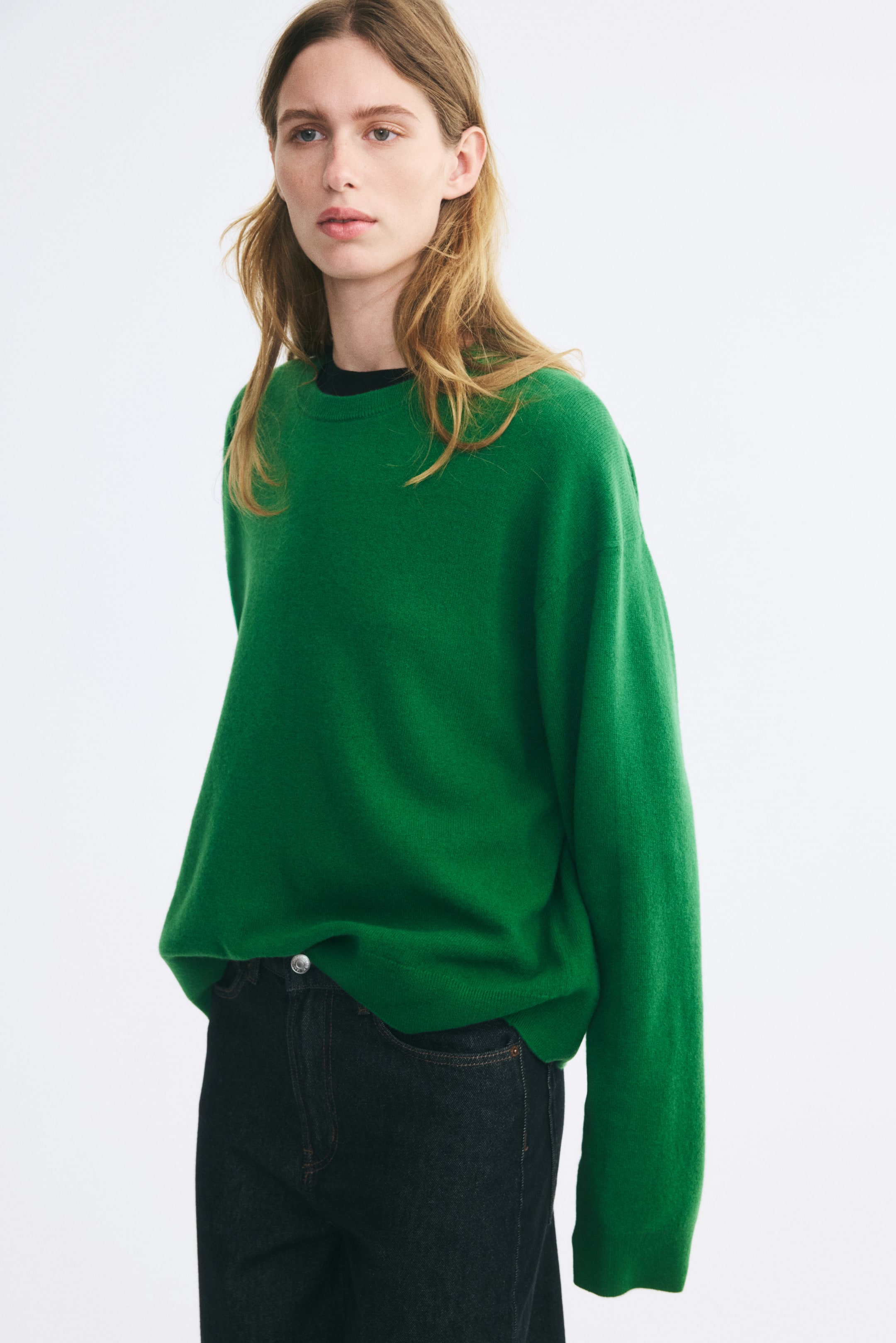 View larger image: An oversized green knitted wool sweater is worn by the model, featuring a round, rib-trimmed neckline, dropped shoulders, and long sleeves. The sweater is layered over a black crew-neck top and partially tucked into dark jeans.