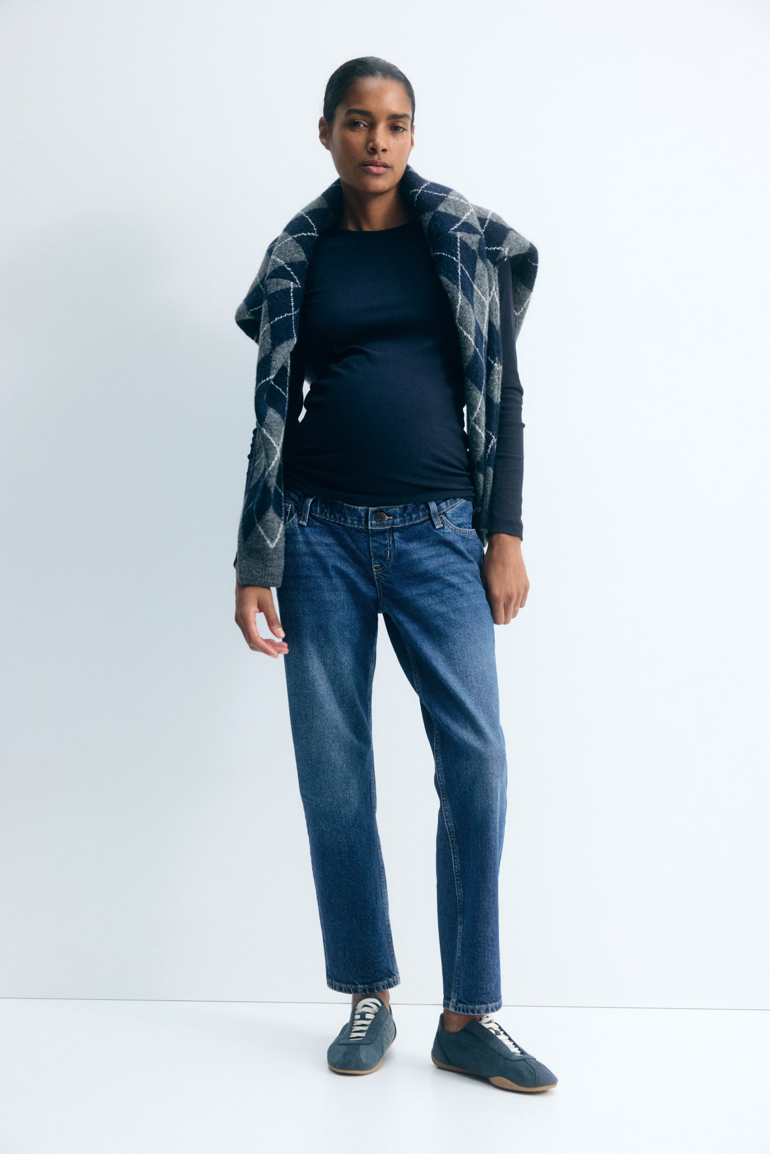 Modelen bÃ¦rer mellemvaskede blÃ¥ denimmammabukser med falsk gylp og knap, stylet med en mÃ¸rk langÃ¦rmet top og blÃ¥ sneakers. En grÃ¥ og marineblÃ¥ argyle-sweater er draperet over modelens skuldre og komplementerer de ankellange jeans med smalle ben. - 1