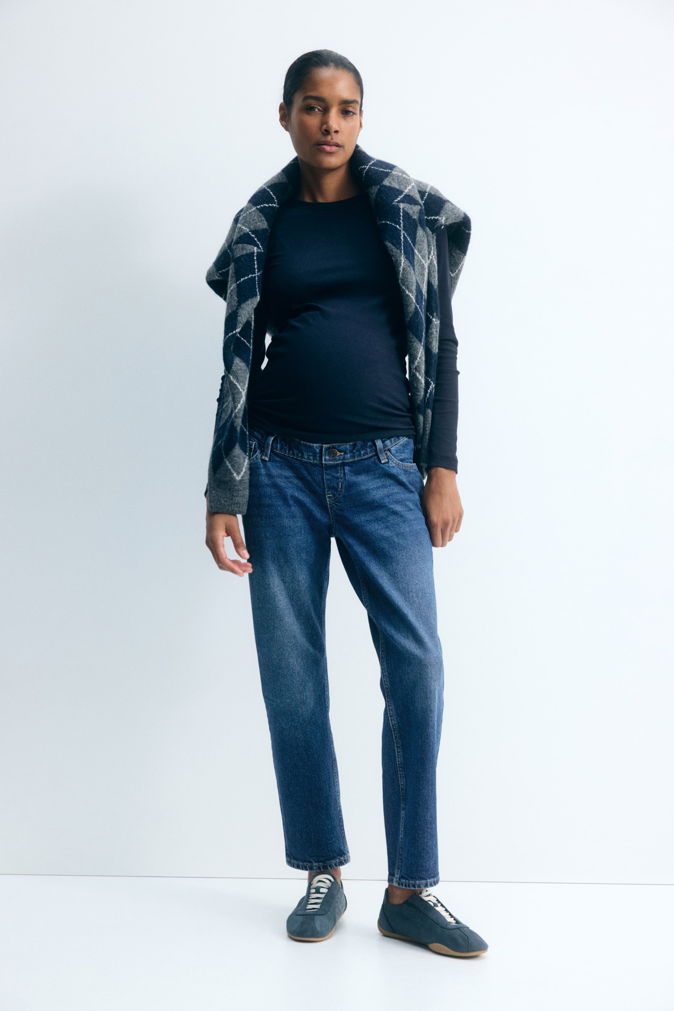 Se større billede: Modelen bærer mellemvaskede blå denimmammabukser med falsk gylp og knap, stylet med en mørk langærmet top og blå sneakers. En grå og marineblå argyle-sweater er draperet over modelens skuldre og komplementerer de ankellange jeans med smalle ben.