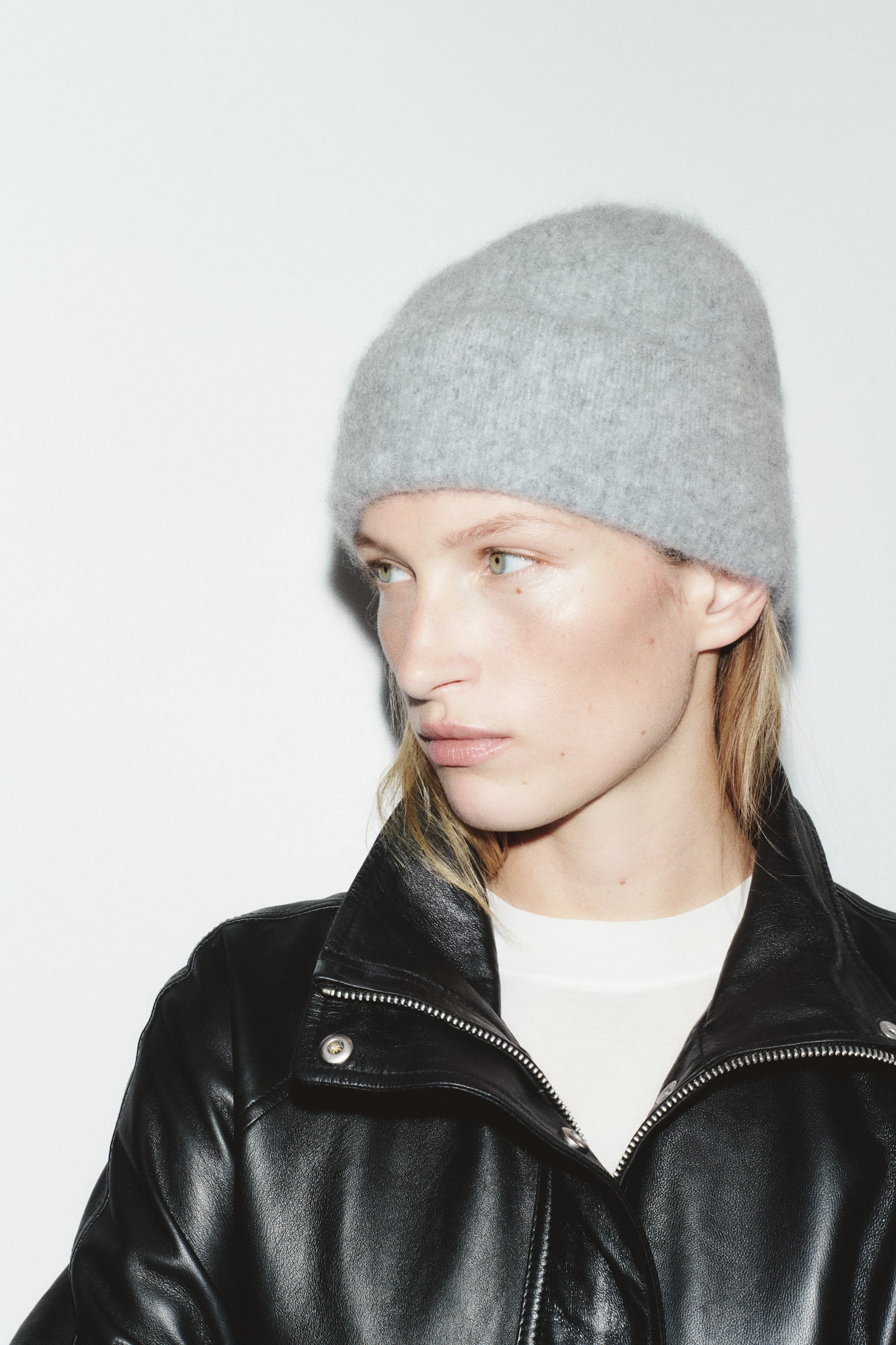 A light grey beanie in a soft, brushed-finish knit with a turn-up hem is being worn by the model. The beanie is styled with a black leather jacket and a white inner top. - 1