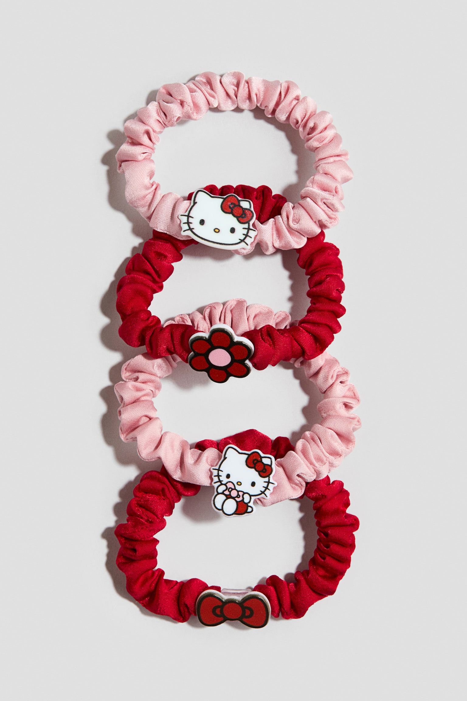 Four gathered satin scrunchies in alternating light pink and deep red are interlinked vertically, displaying their front designs. The pink scrunchies feature Hello Kitty character charms, while the red scrunchies showcase a red flower with a pink centre and a red bow charm.