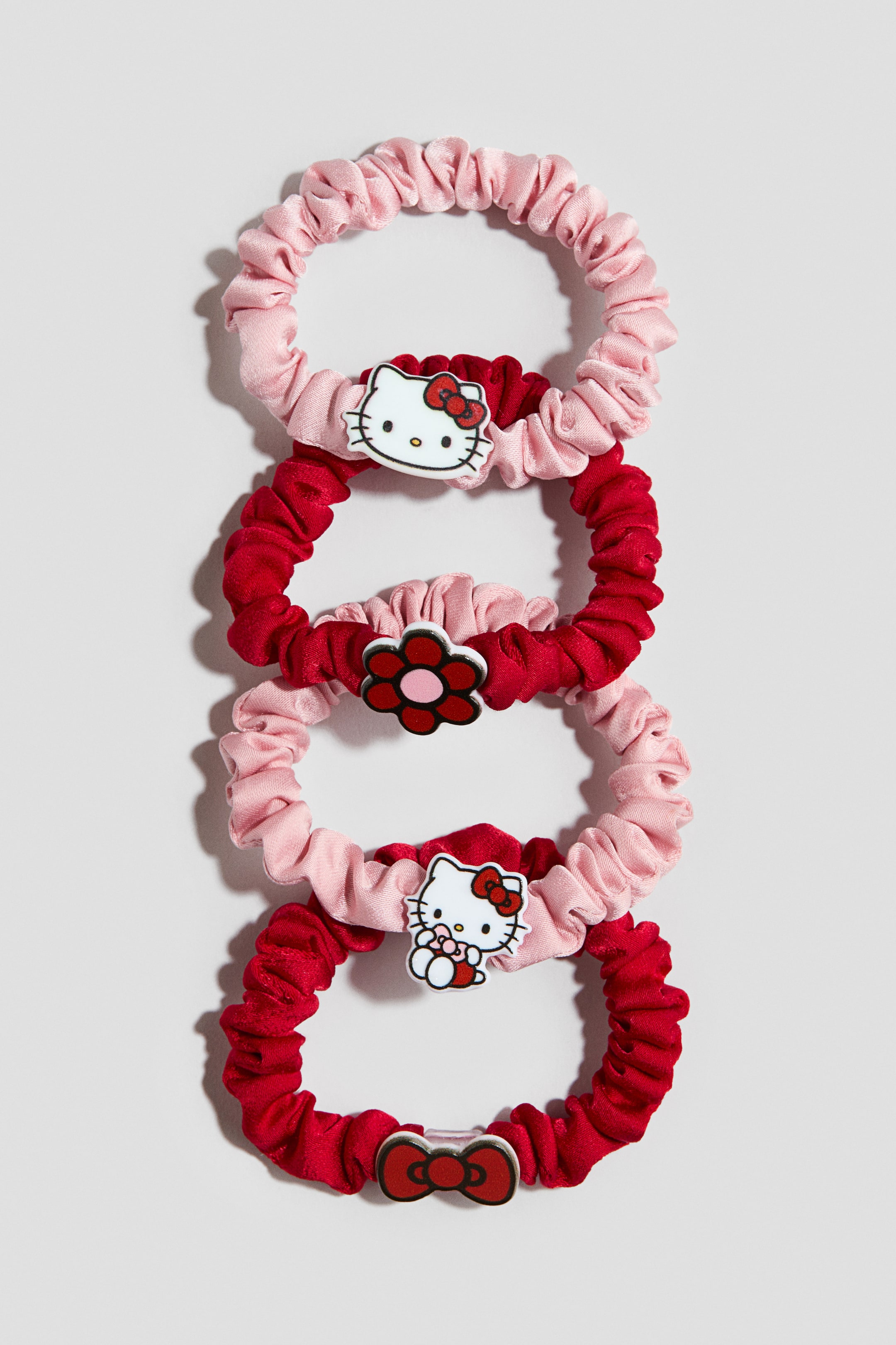 Se større billede: Fire rynkede satinhårbånd i skiftevis lyserød og dyb rød er flettet sammen lodret og viser deres frontdesign. De lyserøde hårbånd er prydet med Hello Kitty-figurcharme, mens de røde hårbånd fremviser en rød blomst med et lyserødt centrum og en rød sløjfecharm.