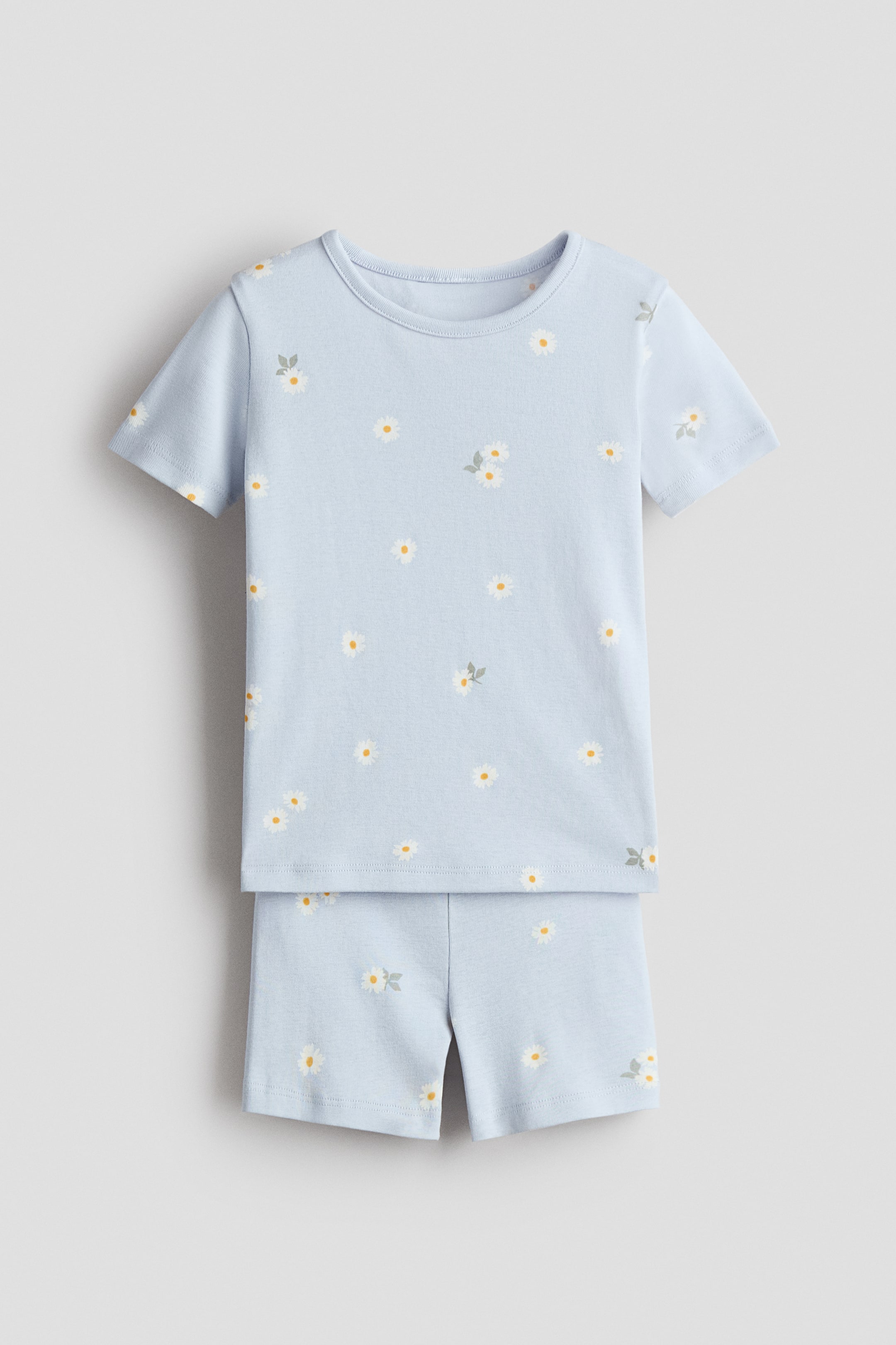 View larger image: A light blue pajama set made from soft jersey fabric is shown from the front. The set consists of a T-shirt with short sleeves and a narrow-trimmed round neckline and matching shorts, both featuring an all-over print of white daisies with yellow centers and small green leaves.