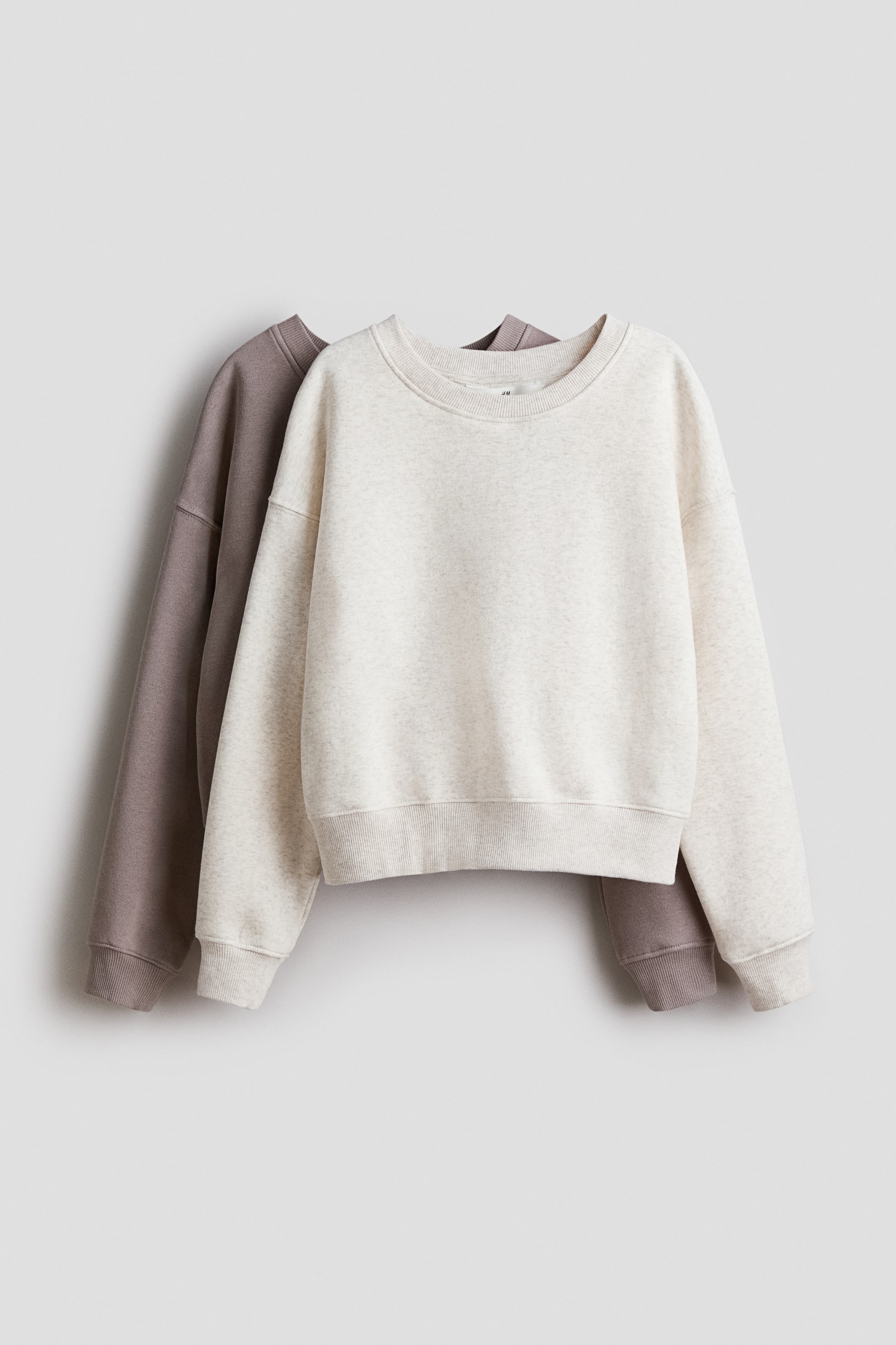 View larger image: A light beige long-sleeved sweatshirt is presenting its front, featuring a round, rib-trimmed neckline, dropped shoulders, and ribbed cuffs and hem. A darker taupe sweatshirt is partially visible behind it.