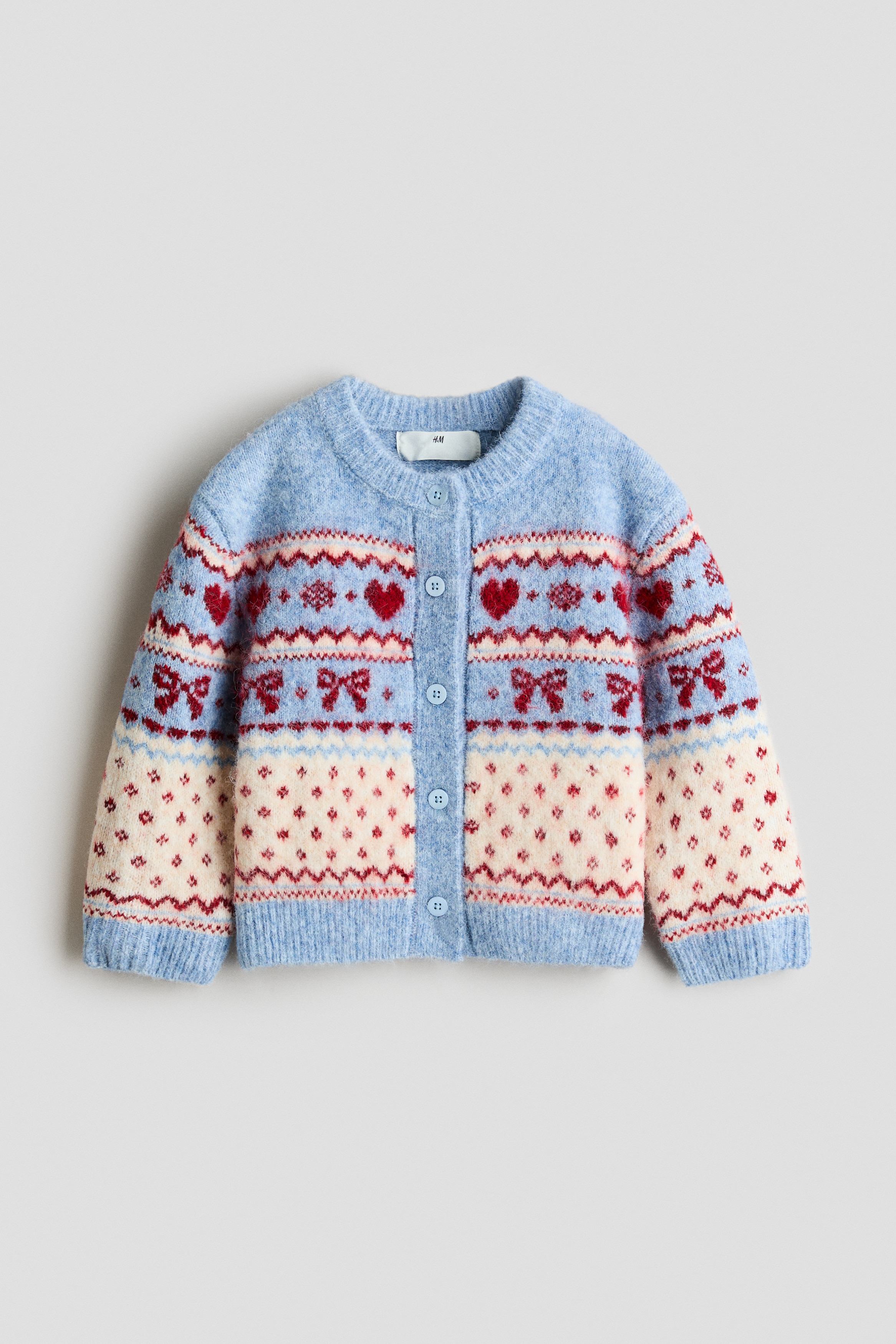 A relaxed-fit jacquard knit cardigan is shown from the front, featuring a pattern of red hearts, bows, and geometric motifs in light blue, cream, and red. The cardigan is designed with a button-down front, dropped shoulders, and ribbed cuffs and hem, styled with a cream knit hood draped over the chest.