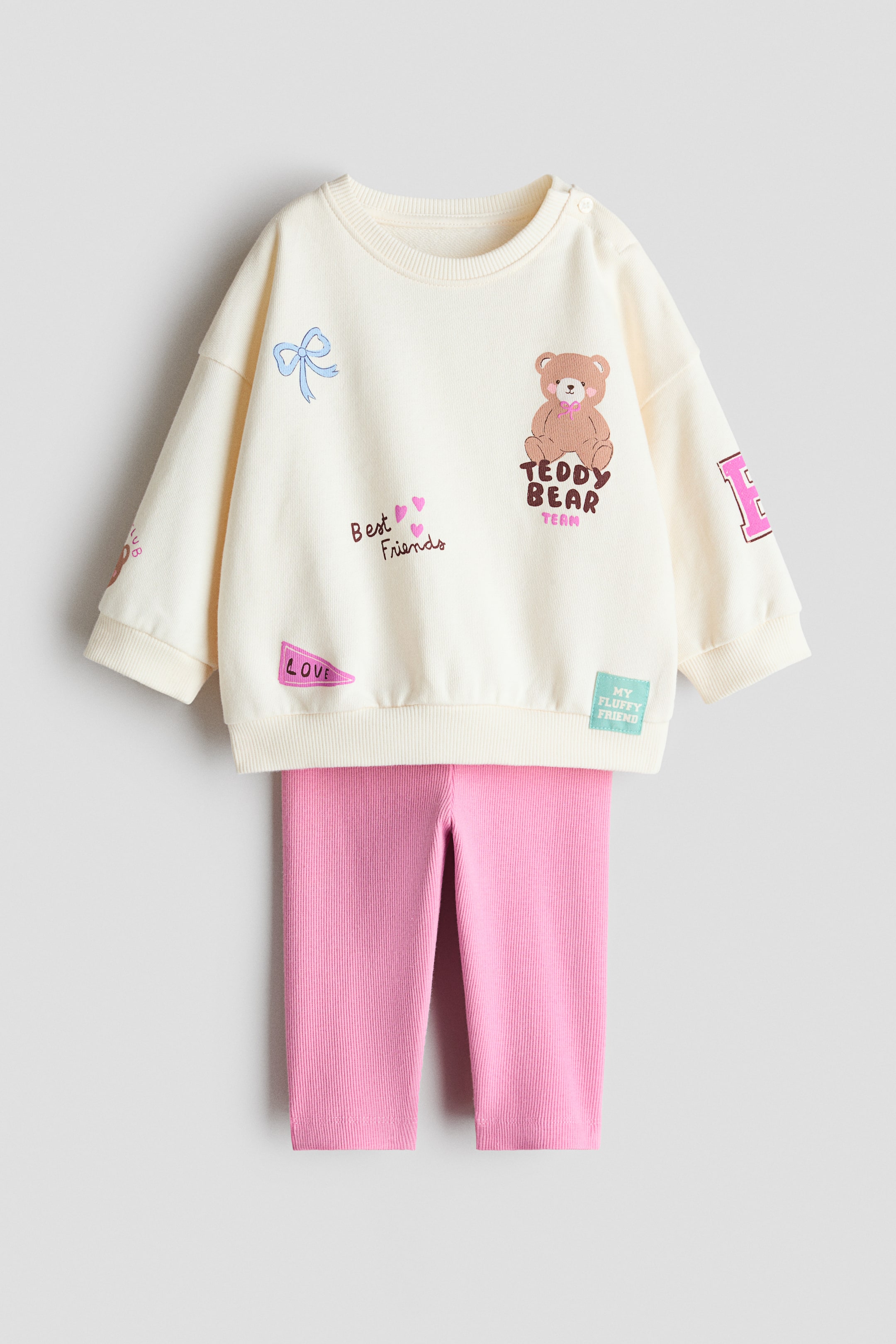 Grösseres Bild ansehen: Ein zweiteiliges Set präsentiert sich mit einem cremefarbenen Sweatshirt-Oberteil, das einen mehrfarbigen Druck mit einem Teddybär, Textelementen und einer Schleife zeigt. Es verfügt über einen runden, gerippten Ausschnitt mit einer Knopföffnung auf der linken Schulter, tief angesetzte Schultern sowie breite, gerippte Bündchen und einen Saum. Dazu gehören rosa gerippte Leggings mit einem elastischen Bund.