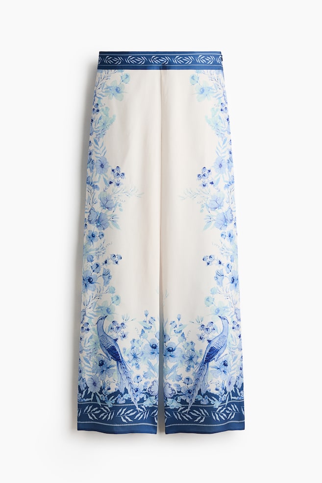 These front-facing, wide-leg pants feature a white base with an intricate blue floral and bird pattern. A dark-blue waistband with a lighter-blue leaf motif sits at the top, and the legs display large blue birds near the hem, which is bordered by a matching dark-blue patterned band. - 1