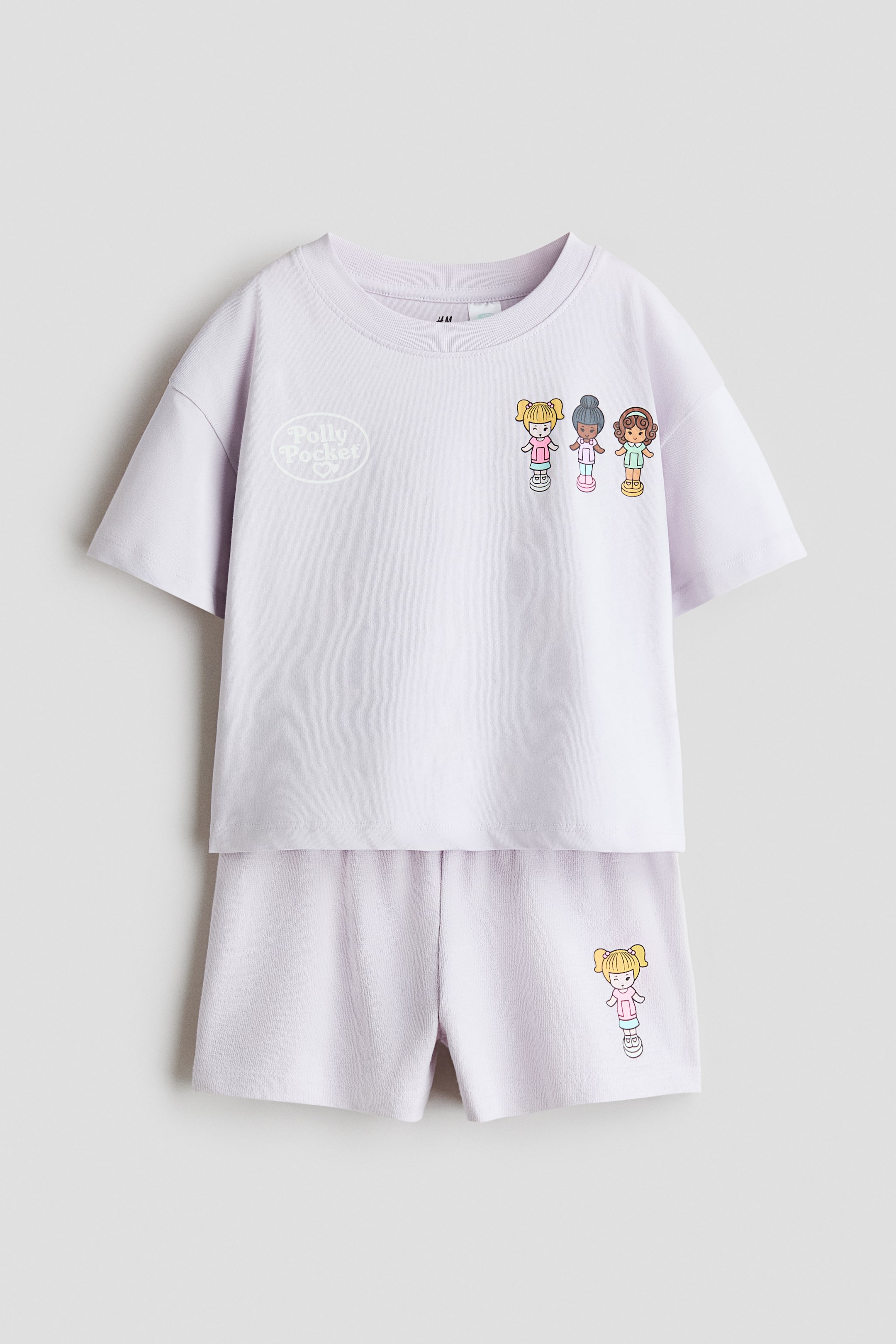 Größeres Bild ansehen: Das Lavendel-Jersey-T-Shirt und Shorts-Set zeigt seine Vorderseite. Das T-Shirt zeichnet sich durch einen runden, gerippten Halsausschnitt, überschnittene Schultern und ein 'Polly Pocket'-Logo zusammen mit drei Charaktergrafiken auf der Brust aus, während die passenden Shorts einen elastischen Bund und eine einzelne Charaktergrafik am linken Bein aufweisen.