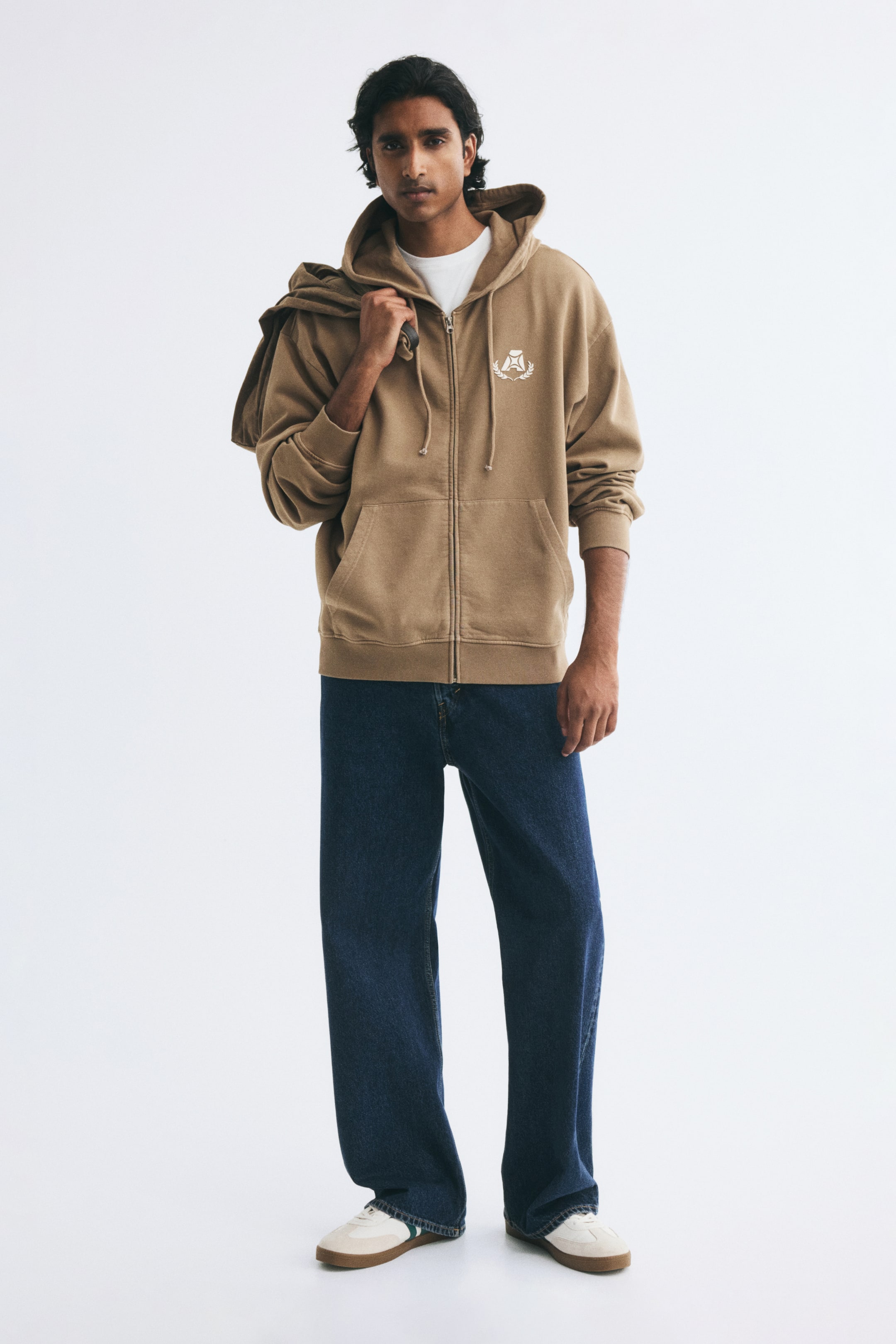 View larger image: A tan zip-up hoodie featuring a small white chest crest, a drawstring hood, and front pockets is being worn by the model over a white T-shirt. The hoodie is styled with dark blue wide-leg jeans, while another tan garment is being carried over the model's shoulder.