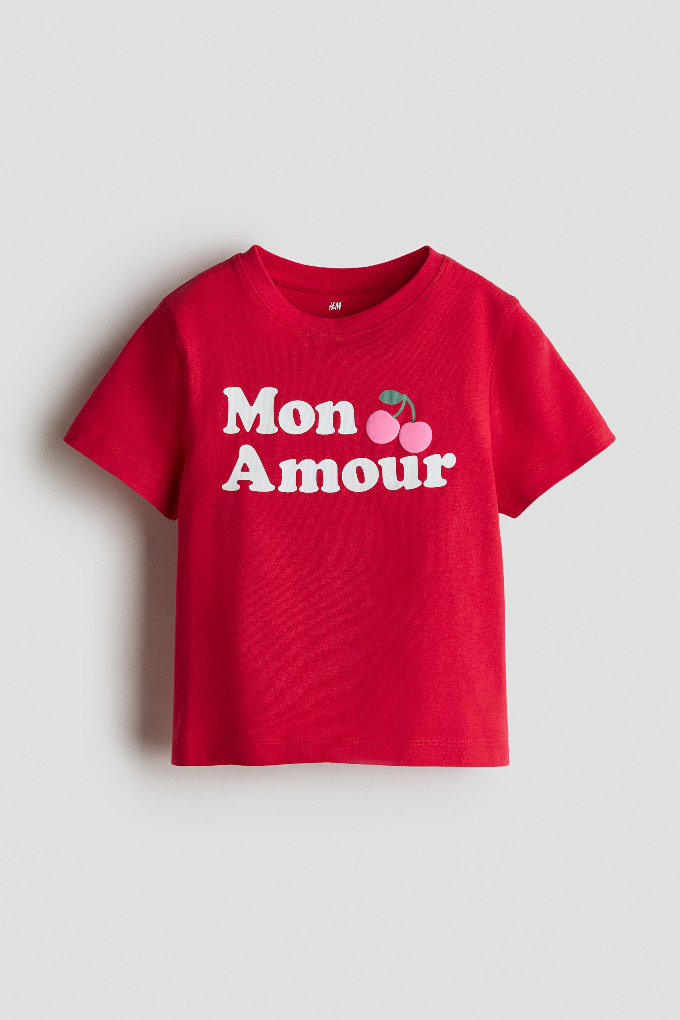 View larger image: A red t-shirt crafted from soft, printed cotton jersey is displayed from the front, showcasing a ribbed trim around the neckline. The front exhibits a print of "Mon Amour" in white lettering accompanied by two pink cherries and green stems.