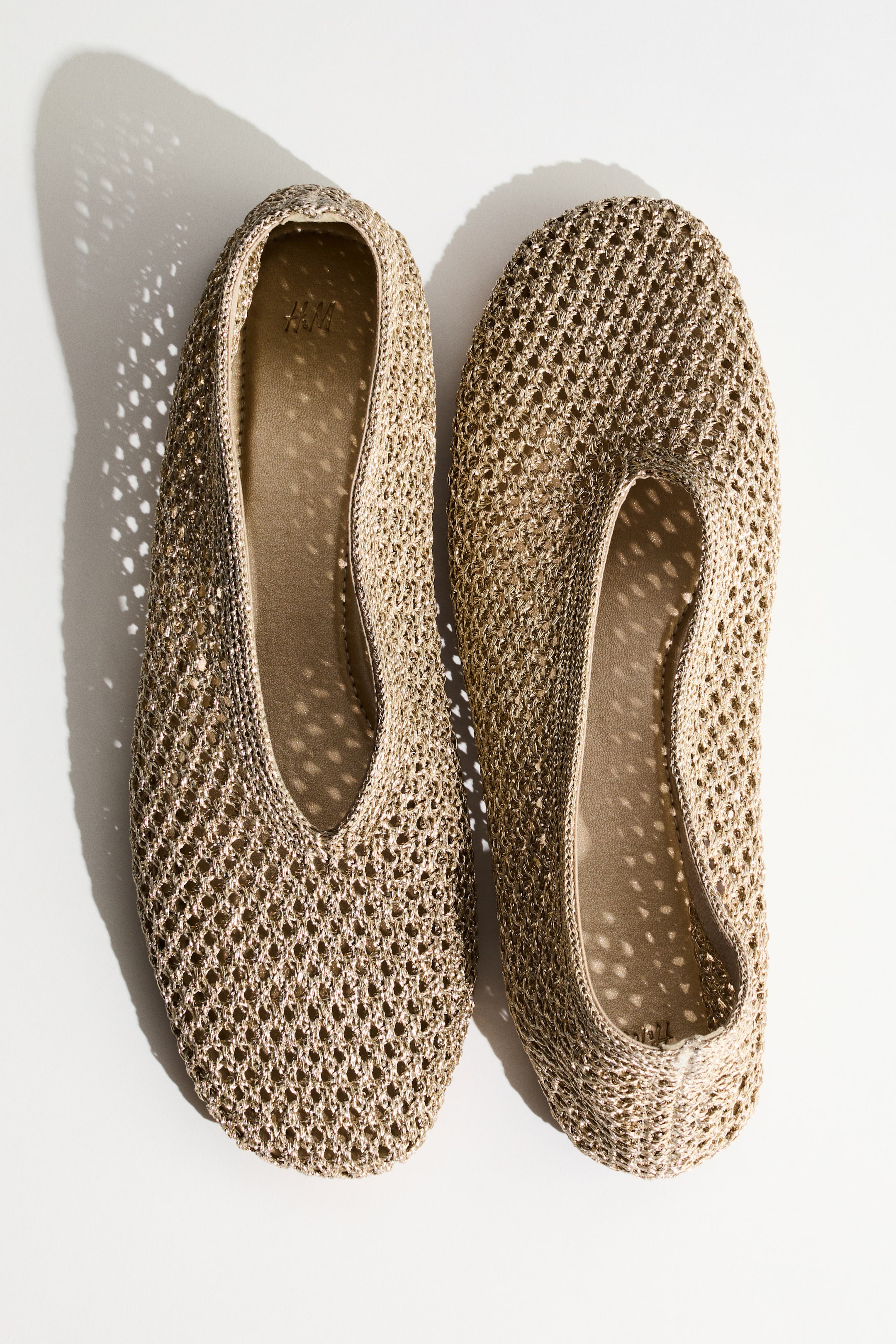 Visa större bild: Ett par gyllene stickade meshballerinaskor visas från en ovanifrån vy. Varje sko visar upp en rundad tå och en synlig beige innersula med ett H&M-logotyp, där den ena skon lutar något åt vänster medan den andra lutar något åt höger.